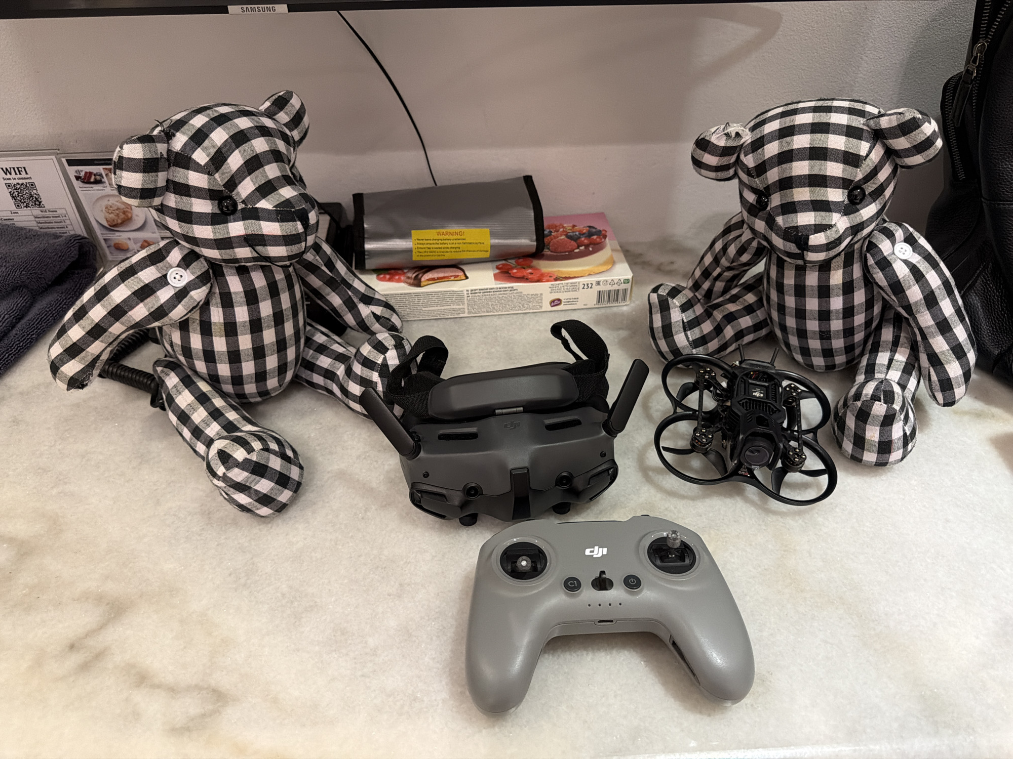 In Ban Phonsavang Neua, Laos, on a mottled white surface, are two black and white checkered teddy bears. Between them, a pair of FPV goggles and a drone controller are laid out. To the left of the goggles is a small black drone. Behind the drone and goggles are a gray fabric pouch with a yellow warning label and a box of pastries. A black backpack is visible on the far right.