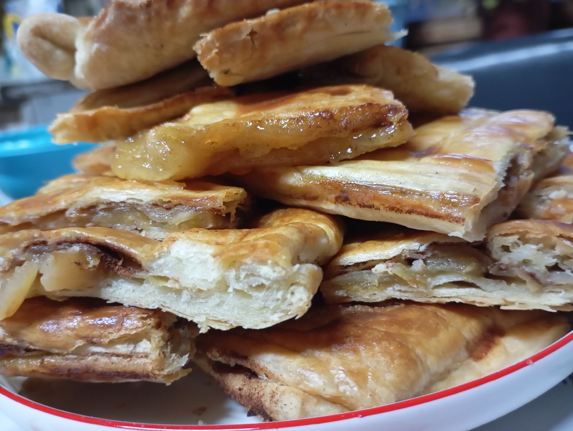 A stack of sliced apple pastries is presented in a shallow white bowl with a red rim. The pastries, made with a flaky, golden-brown dough, are layered with a glistening, cooked apple filling that appears to be spiced. The apples have a soft, translucent quality, and some have a slightly browned edge. The pastries are cut into rectangular pieces and arranged in a somewhat haphazard pile, suggesting they have been served from a larger batch. The lighting is warm and somewhat diffuse, highlighting the textures and glossy sheen of the pastries. The background is softly blurred, but hints of a domestic setting are visible, with a blue bowl in the upper left corner and indistinct shapes in the background that could be kitchen elements. There are no people or animals visible in the image. The scene depicts a food item, likely a dessert or snack, presented in a home environment. The image does not provide any specific geographical indicators that would allow for pinpointing its location within the city of Фоки, Russia.