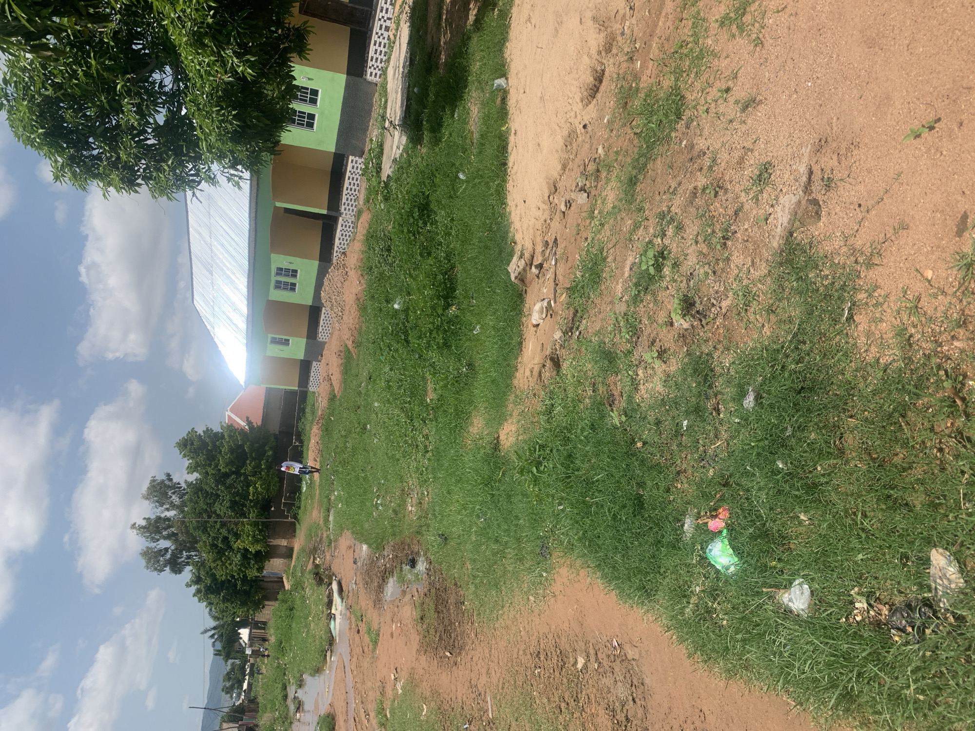 The image depicts a street scene in Jalingo, Nigeria. A dirt road runs diagonally across the foreground, with a muddy, waterlogged area to the left and a grassy verge to the right. The grassy verge has litter scattered within it, including plastic bags. In the mid-ground, a person is walking on the dirt road. Beyond the road, a building with a metal roof and painted walls in shades of yellow, green, and black stands. The building has multiple windows and an overhang. Trees are visible behind and to the side of the building, and in the distance, hills can be seen under a partly cloudy sky. The overall impression is of a developing area with some signs of neglect.