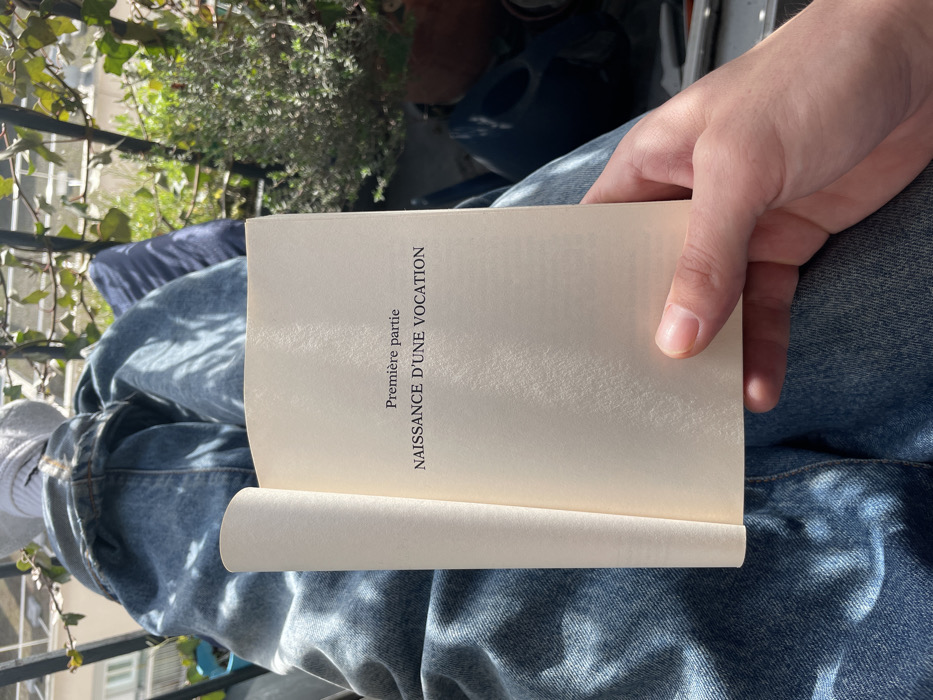 A first-person perspective image depicts an individual seated on a balcony in Paris, France, with their legs extended. The subject is wearing blue denim jeans and white socks. Their right hand holds open a cream-paged book, displaying the French text "Première partie" and "NAISSANCE D'UNE VOCATION". In the background, a black metal railing is visible, adorned with green foliage including a potted plant in a terracotta pot on the right. A blurred building appears in the far distance, under bright sunlight, indicating an outdoor daytime scene.