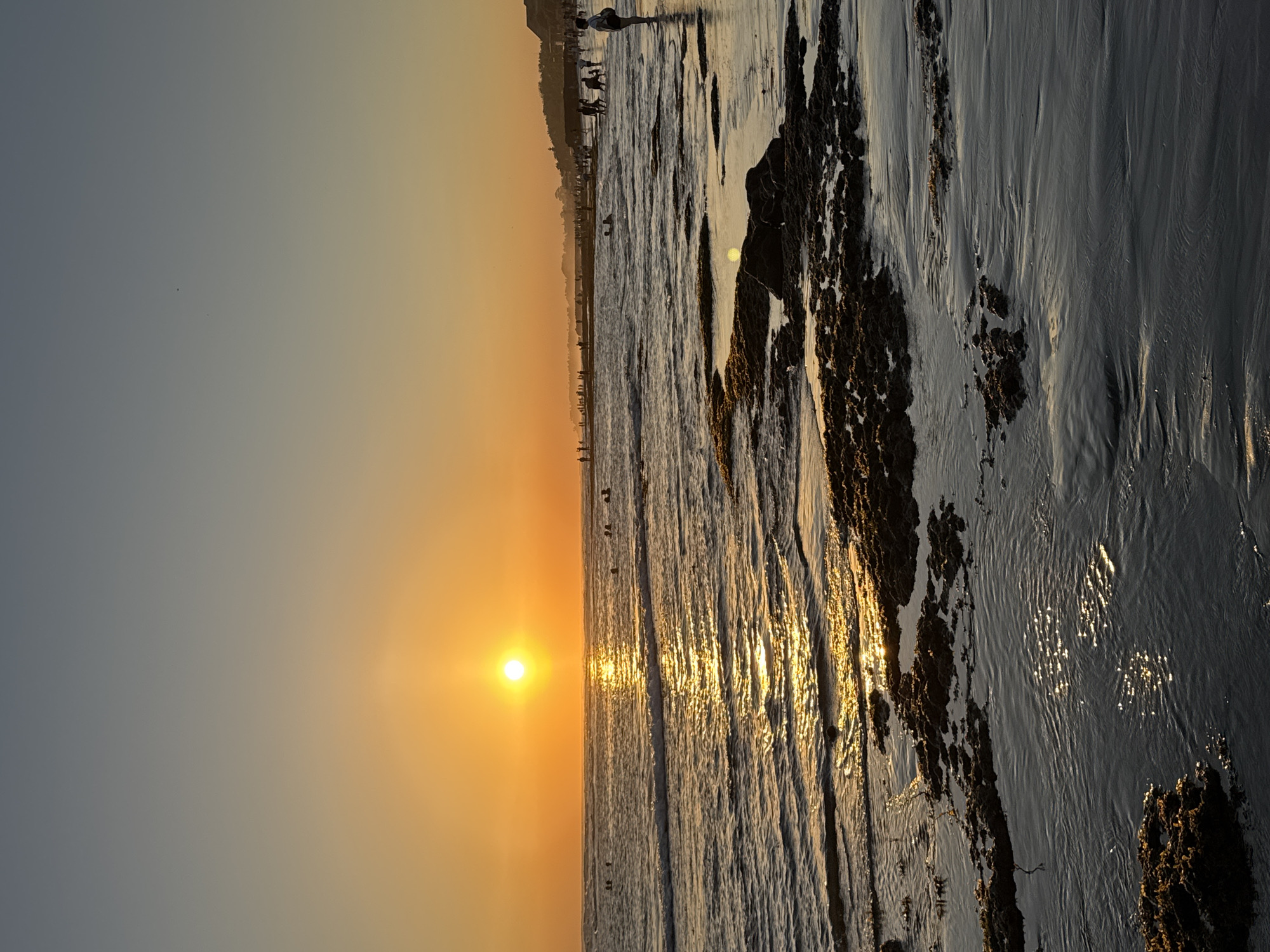 This media file captures a sunset scene at a beach in North Kuta, Indonesia. The image, oriented vertically, shows a wide expanse of ocean meeting a sandy shore. The sun, a bright orange orb, is low on the horizon, casting a warm glow across the sky and reflecting shimmering golden light on the water's surface. The sea appears calm, with gentle waves lapping at the shore.
Along the waterline and further up the beach, numerous people can be seen. Some are walking along the edge of the water, while others appear to be gathered in groups. Their silhouettes are visible against the bright sunset, suggesting a leisurely evening at the beach. The sand itself shows textures from the receding tide, with patches of darker, wet sand and areas of dry, lighter sand. In the foreground, the beach is punctuated by dark, rocky formations that rise out of the sand and are partially covered by water. The overall mood of the scene is peaceful and idyllic, characteristic of a serene sunset at a popular coastal location. The time of day is clearly evening, as indicated by the low angle of the sun and the warm, diffused light. There is no visible text in the image.