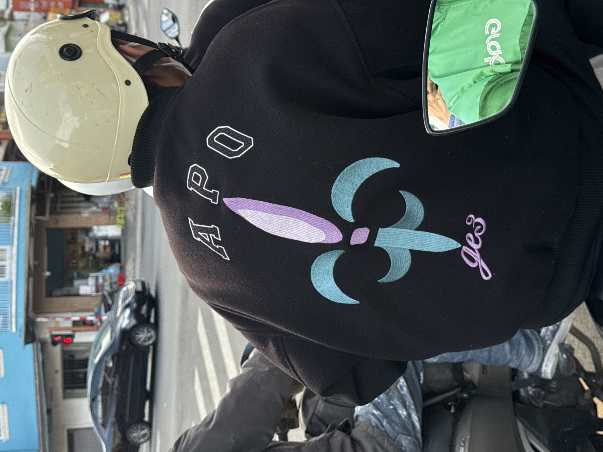 The image shows a person from the back, wearing a black jacket with "APO" embroidered in white on the upper back and a fleur-de-lis design in purple and teal below it, with the word "jes" embroidered in purple script at the bottom. The person is wearing a light-colored helmet. They are sitting on a motorbike on a street, indicated by the crosswalk lines on the road and a black car in the background. The environment suggests a city setting with buildings and streetlights. The reflection in the mirror of the motorbike shows another person wearing a bright green jacket with white lettering, possibly a uniform. The overall scene is consistent with urban street life in Da Lat, Vietnam, which is known for its motorbikes and busy streets.