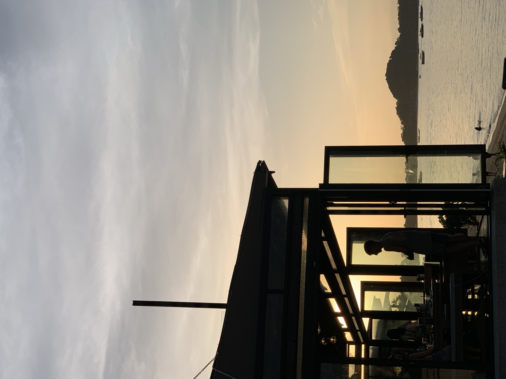 The image depicts a serene scene at dusk on the island of Ko Samui, Thailand. A silhouetted open-air restaurant with a dark roof and glass doors overlooks a calm body of water. Inside the restaurant, the silhouettes of several people are visible, some seated at tables, others standing. The exterior lighting of the restaurant casts a warm glow through the glass.
Outside, the sun is setting, casting a soft orange and yellow hue across the sky and water. In the distance, a lush, tree-covered island rises from the sea, with several small boats anchored offshore. The water's surface is gently rippled, reflecting the colors of the sky. A few people are visible near the water's edge, with one person appearing to swim. The overall atmosphere is tranquil and picturesque, capturing the essence of a tropical evening.