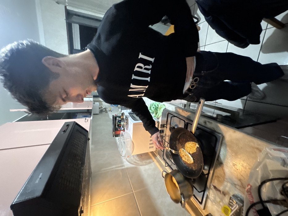 This image depicts an indoor kitchen scene in the city of Bragança, Portugal. A young adult male is standing at a gas stovetop, actively engaged in cooking. He possesses a light skin tone and dark hair, and is dressed in a black long-sleeve sweatshirt featuring "AMIRI" in white lettering across the chest, dark blue jeans, and a dark belt with a gold-colored double-C shaped buckle. His white underwear is visible above the waistband of his jeans.
The male subject holds a silver-colored fork in his right hand, using it to manipulate two pieces of light-colored, possibly breaded, food within a black frying pan situated on the front-right burner of a four-burner gas stove. A silver pot with a lid handle is positioned on the rear-left burner, containing a yellowish substance.
The kitchen environment includes a light-colored countertop, likely marble or granite, where a white plastic bag and other items are visible on the lower left. A black range hood with controls is mounted above the stovetop. Above the range hood, pinkish-brown overhead cabinets are present. A tiled backsplash covers the wall behind the stove. To the right, a white microwave oven rests on the countertop, topped with a wire rack. A light green cloth is situated near the microwave. Artificial lighting illuminates the area, casting light on the cooking surface. A window or door with a dark exterior is visible in the background, suggesting evening hours.
