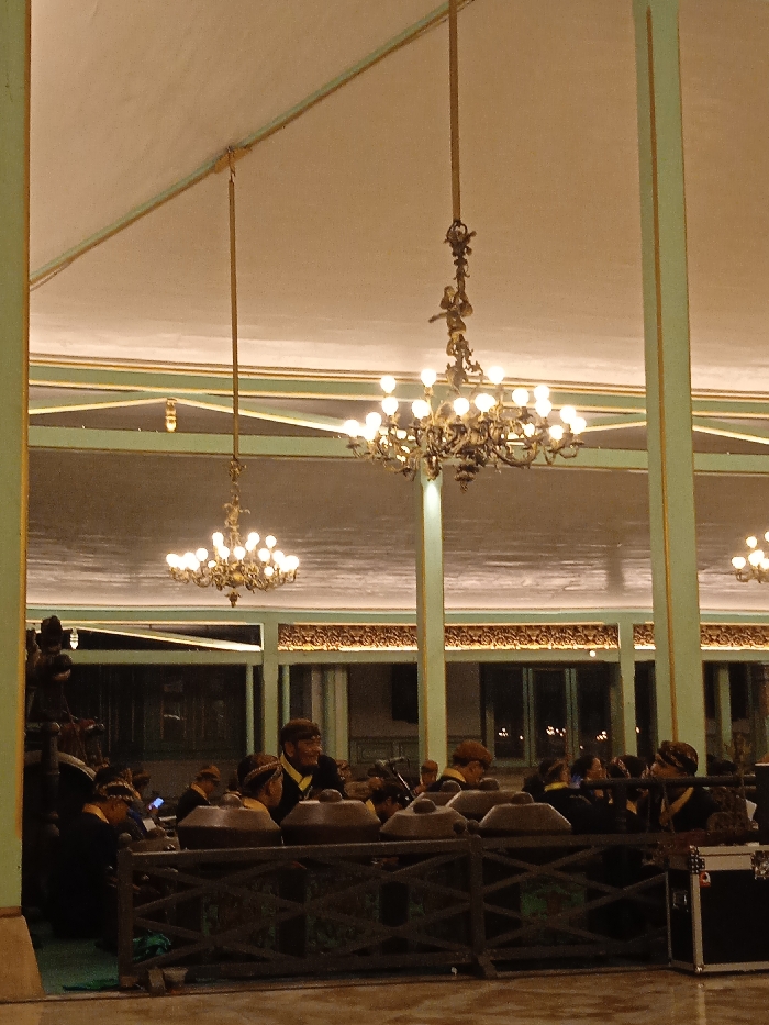 The image shows a large room with high ceilings and a light-colored tiled floor. Two ornate chandeliers with incandescent bulbs hang from the ceiling. There are several people sitting behind a wooden railing, wearing traditional clothing and hats. There is a large black speaker box sitting in the front corner of the room, near the railing. The scene is taken in a large building located at -7.559, 110.783 in Surakarta, Indonesia. This location is within the grounds of the Kraton Kasunanan Surakarta, the palace of the Surakarta Sunanate. The room appears to be a grand hall, perhaps a reception area or a performance space.