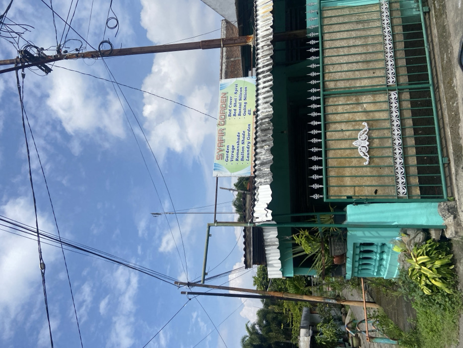 The image shows a storefront in Mojokerto, Indonesia, located at latitude -7.458 and longitude 112.446. The store, painted a vibrant turquoise, is situated on a narrow street and features a sign reading "SYAFIR GORDEN". The sign lists various products sold, including curtains, bed covers, bed sheets, and bedding. A green metal gate with intricate details separates the store from the street, and multiple power lines crisscross the sky. Lush greenery can be seen surrounding the store and on the opposite side of the street, while the sky is a bright blue with scattered white clouds.