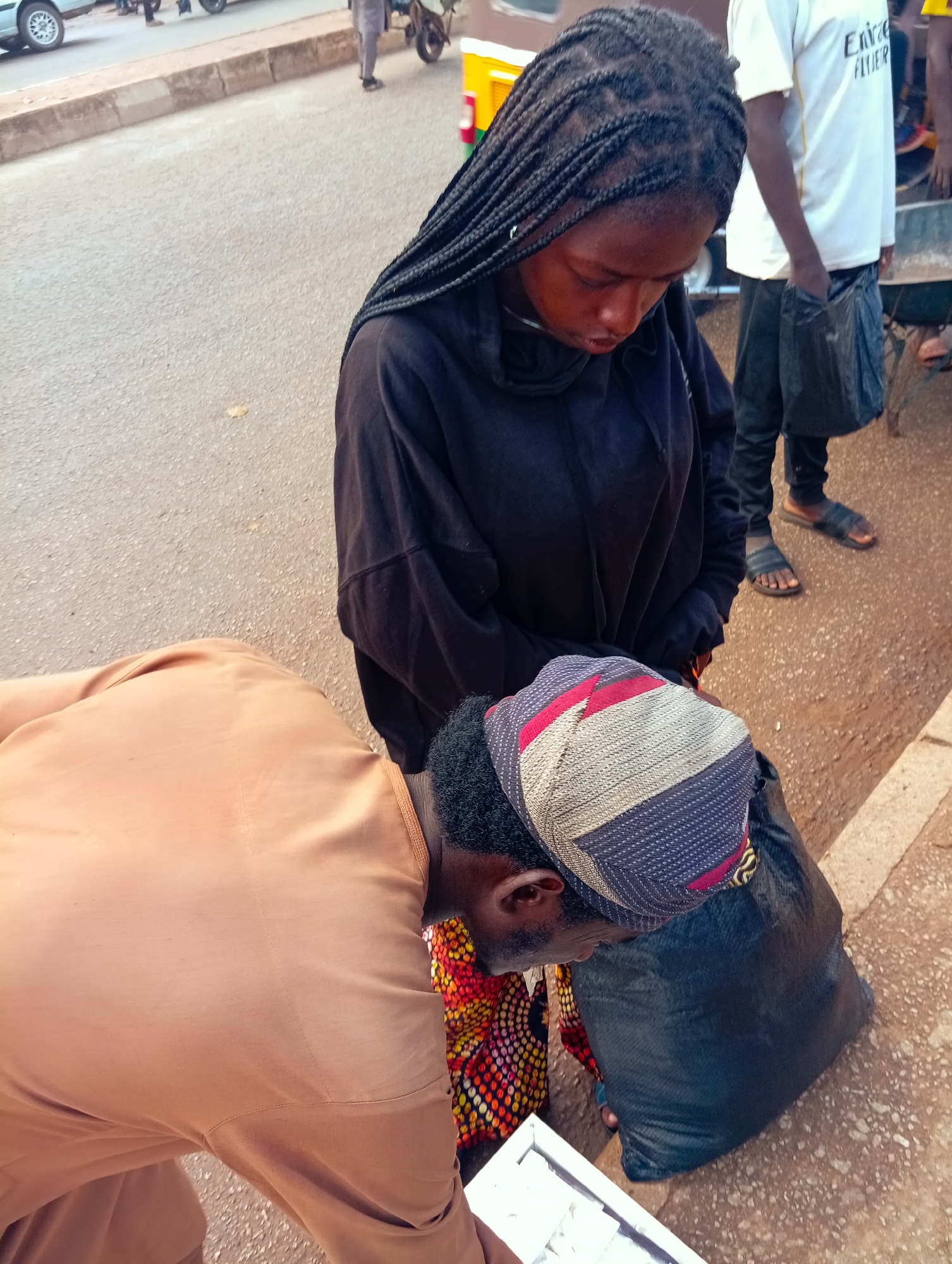 In Bukuru, Nigeria, two individuals are captured in an outdoor scene on what appears to be a street. The foreground is dominated by a person wearing a tan outfit, bending down. They have dark, curly hair visible at the nape of their neck. This individual is looking towards a white object held in their hands.
Behind them, another person, a young woman, is visible. She has dark, braided hair that cascades down her back. She is wearing a black hooded garment and is also looking downwards, though her focus is not on the white object. On her head, she wears a headwrap with a distinctive pattern of red, white, and blue. Next to her, and slightly behind, is a large black plastic bag.
The background reveals more of the setting. A paved surface, likely a road, stretches into the distance, with a raised curb or sidewalk. A yellow and green vehicle, possibly a tuk-tuk, is partially visible. In the further background, other vehicles and people are present, suggesting a public area or marketplace. The lighting suggests it is daytime, and the overall atmosphere appears to be one of everyday activity. There is no visible text in the image.