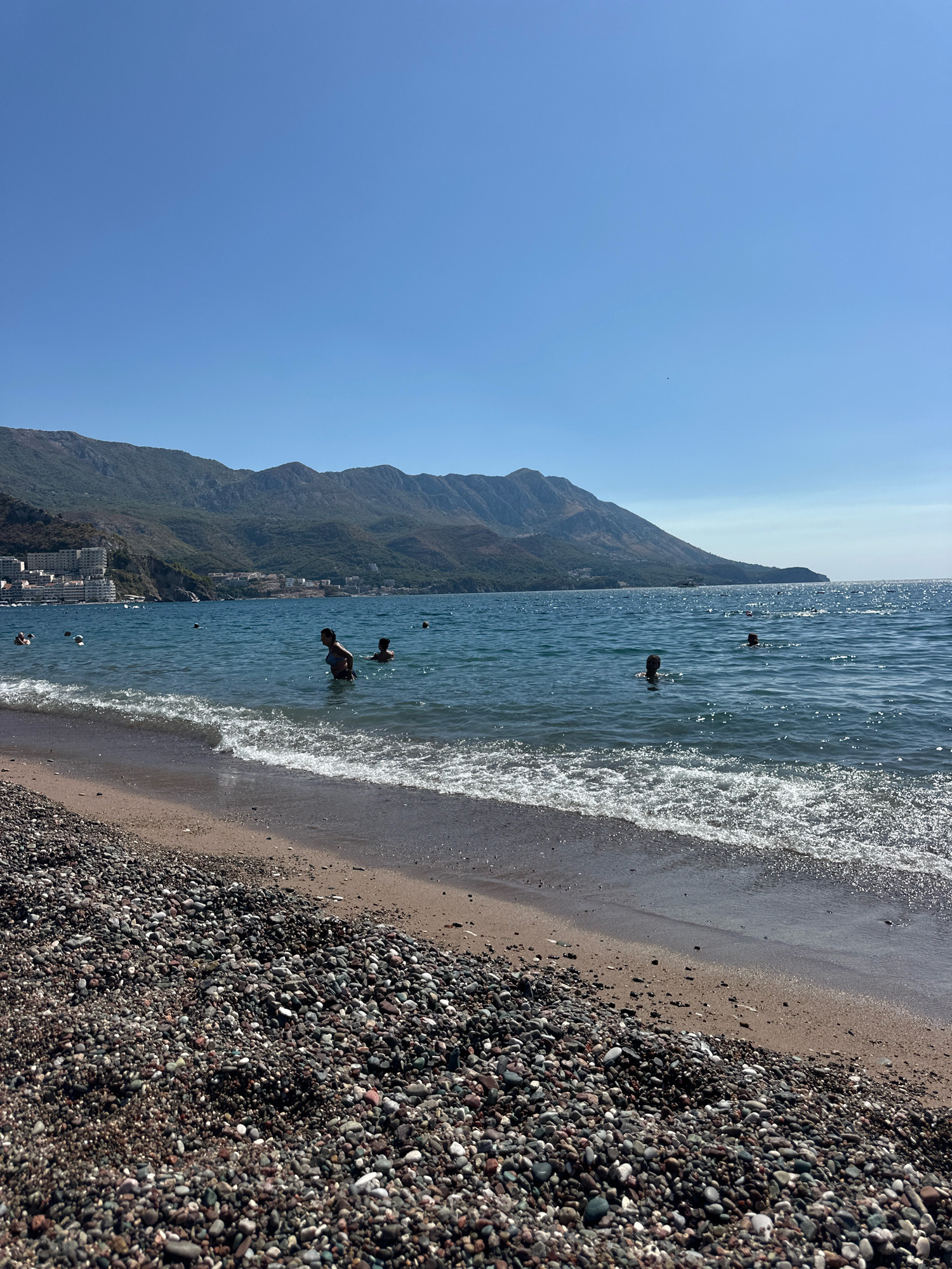 A wide shot captures a pebble beach in Boreti, Montenegro, with the Adriatic Sea and a mountainous coastline under a clear blue sky. Several people are swimming and wading in the shimmering blue water, with some in the foreground and others further out. In the distance, buildings line the coast nestled against the verdant green mountains. The foreground is dominated by the textured expanse of the pebble beach, meeting the gentle waves of the sea. The sun glints off the water, creating a sparkling effect.