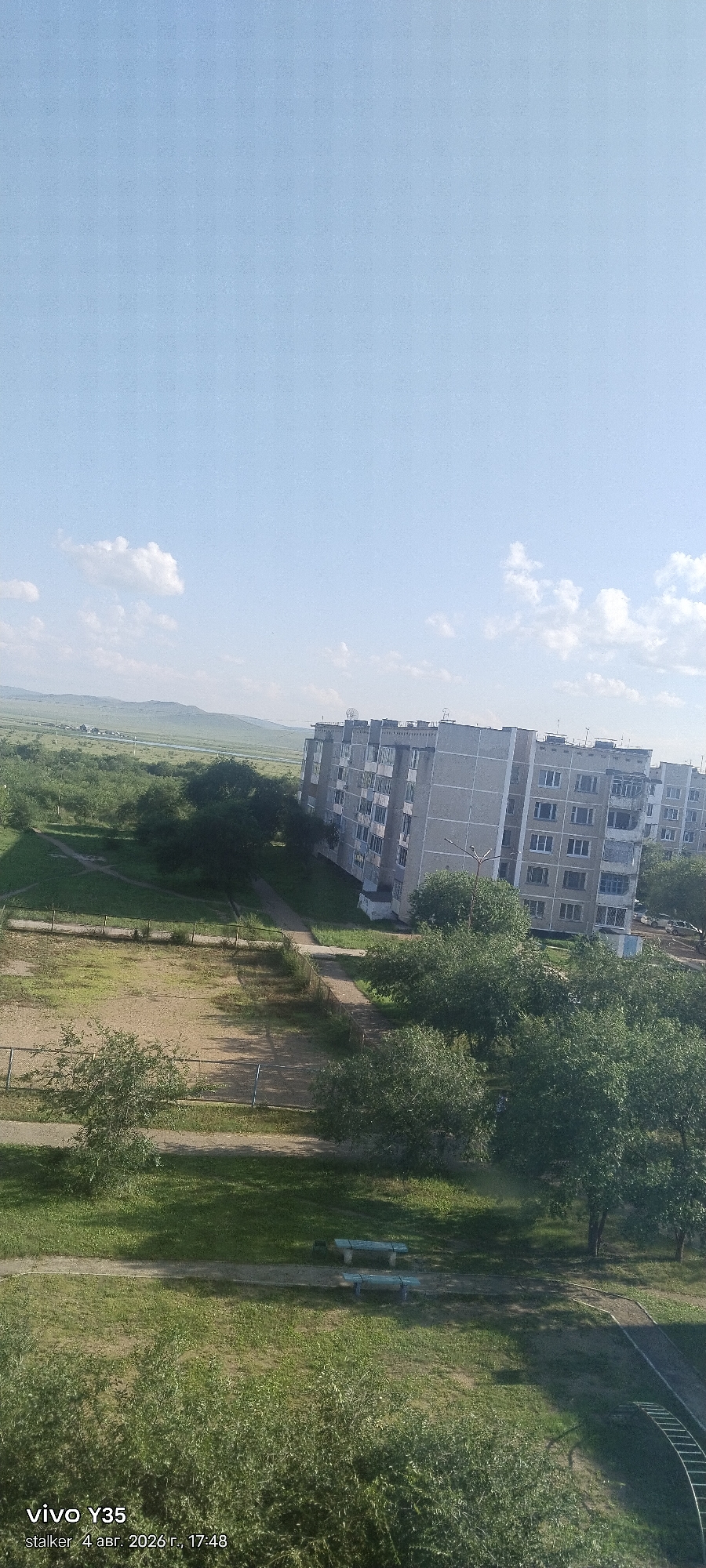 The image depicts a daylight scene in Krasnokamensk, Russia. In the foreground, there are various trees and a dirt path leading to a small playground with a metal climbing structure. Further into the scene, there are two park benches on a grassy area. A large, multi-story residential building with a grey facade and many windows is prominently featured. The building appears to be several stories high and has a modern architectural style. Beyond the building, there are more trees and a vast, open landscape with rolling hills and a clear blue sky with scattered clouds. The overall impression is one of a residential area in a semi-rural or suburban setting. The timestamp at the bottom left indicates "vivo Y35 stalker 4 abr. 2026 r., 17:48".
