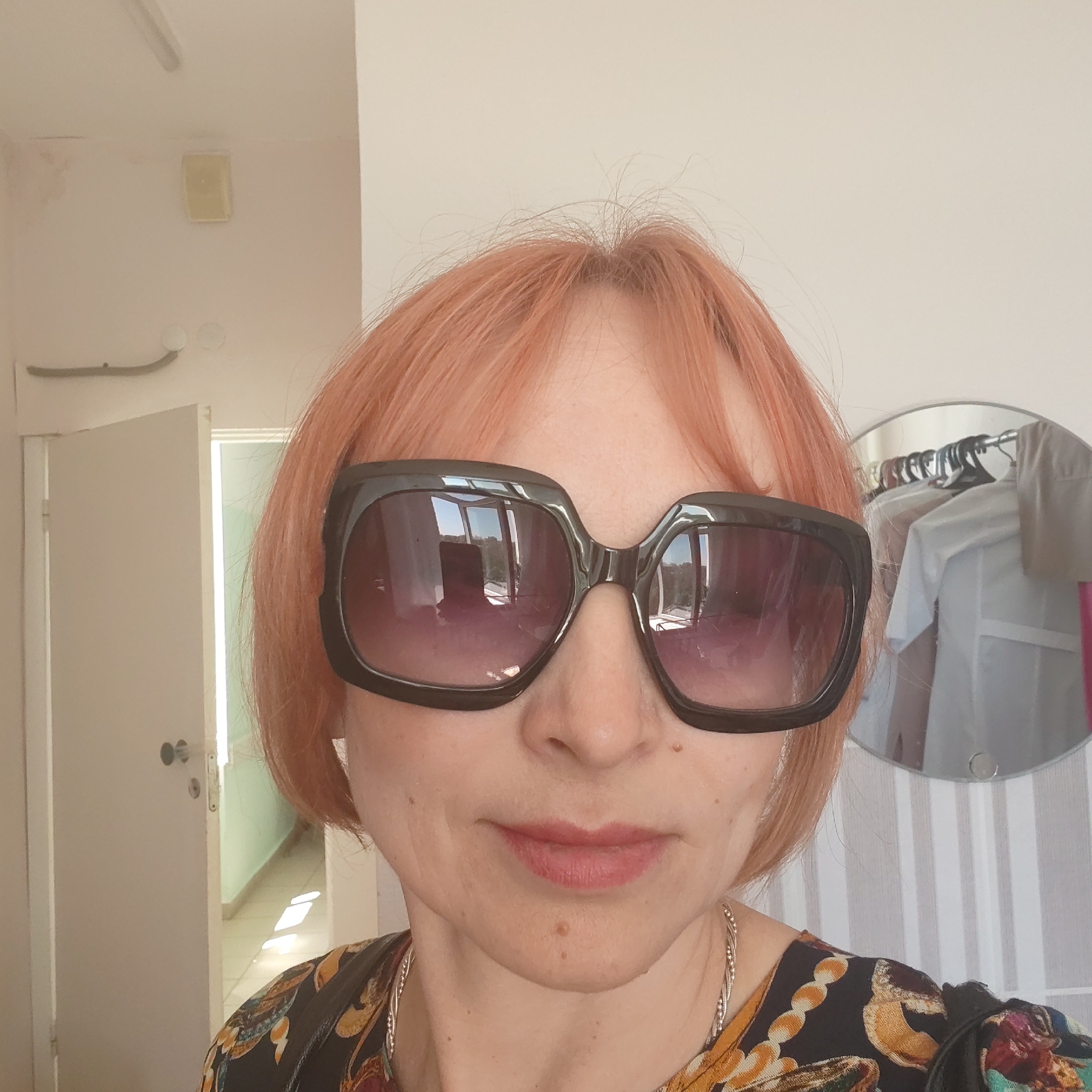 A woman with peach-colored, short, straight hair is posing for a selfie indoors. She is wearing oversized, dark-framed sunglasses. The glasses reflect a windowed area with trees. She has fair skin and is wearing a dark, patterned top with a silver necklace.
Behind her is a white interior with doors, a round mirror reflecting clothes hanging on a rack, and other interior fixtures.
The location of the image is not determined.