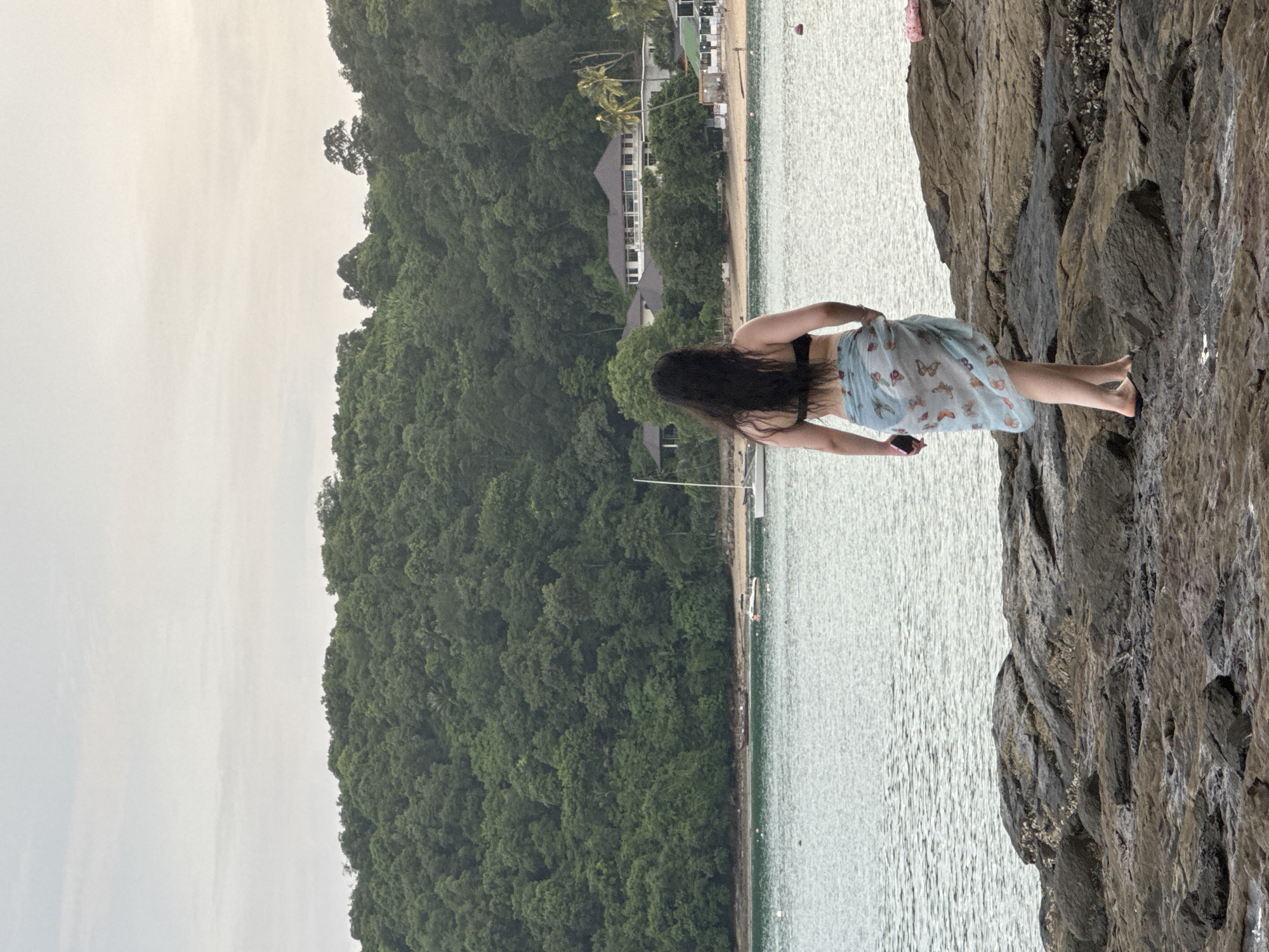 A woman stands on a rocky shore facing a body of water with a dense, green hillside behind it. The woman has long, dark hair and is wearing a black bikini top and a light blue sarong patterned with butterflies. She is holding a pink smartphone in her left hand. The water is calm with a few buoys visible. In the background, a sandy beach leads to a line of buildings, including a large, white resort-style structure with a dark roof, surrounded by palm trees and lush vegetation. A sailboat and a small motorboat are anchored in the water. The sky is overcast. The scene appears to be a coastal resort area, likely in Wichit, Thailand, given the tropical vegetation and resort architecture.