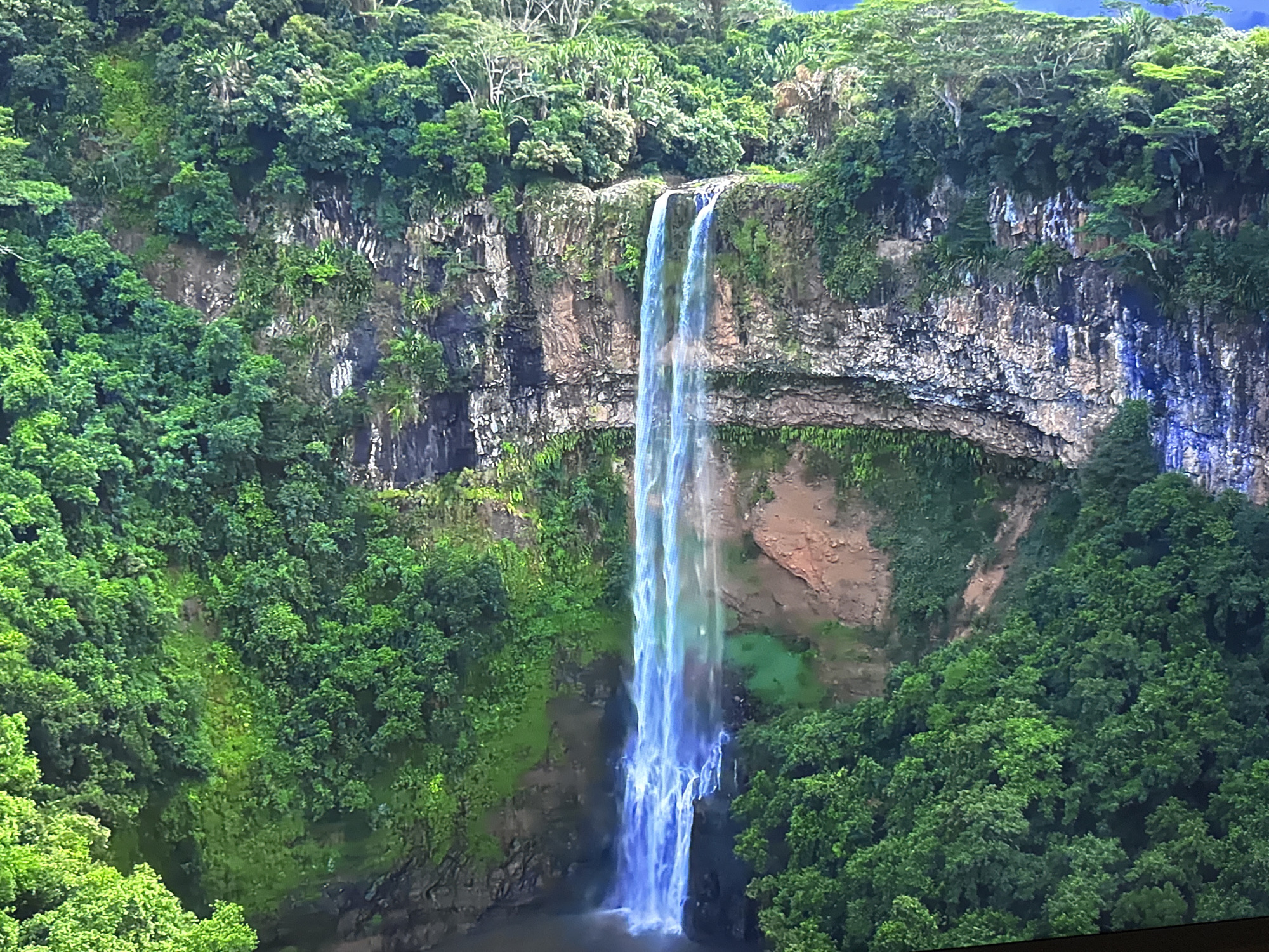 The image shows a tall, narrow waterfall cascading down a rocky cliff face. The cliff is covered in lush green vegetation, including trees and moss. The waterfall is a bright, almost ethereal blue and white, and it plunges into a dark pool at the bottom. The surrounding environment is dense with trees and foliage, suggesting a tropical or subtropical climate. There are no people or man-made objects visible in the image. The scene is a natural landscape, likely a protected area or park, given the pristine condition of the waterfall and surrounding nature. The specific location in Burgas, Bulgaria, is not identifiable from the image alone.