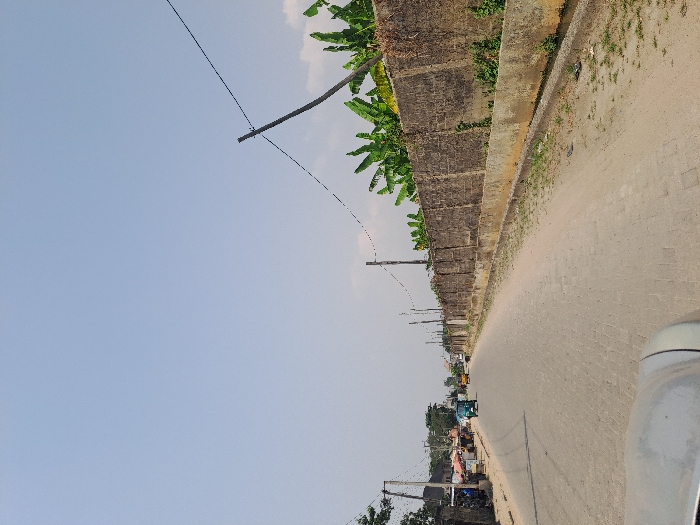 The image depicts a straight road in Badore, Nigeria, under a clear, light blue sky with minimal cloud cover, suggesting bright daylight. The perspective is from the front of a vehicle, with its silver-grey hood visible in the bottom foreground.
The road is paved with interlocking blocks or cobblestones and extends into the distance, narrowing slightly. A dirt shoulder flanks both sides of the paved surface.
On the right side of the road, a long, weathered concrete block wall, approximately 2.5 meters high, runs parallel to the street. Above this wall, dense green foliage, identifiable as large banana leaves, is prominent. A shallow concrete drainage ditch separates the dirt shoulder from the base of the wall. Several wooden utility poles, strung with electrical or communication wires, are positioned along this side.
On the left side of the road, further down the street, various small commercial structures, likely shops or stalls with corrugated metal roofs and awnings, line the roadside. A small group of individuals is visible near these establishments. Additional utility poles with overhead wires are present on this side.
Vehicles are present on the road. A green tricycle (Keke Napep) is visible on the paved surface, traveling away from the camera. A yellow vehicle is further in the background. No specific interactions between individuals are clearly discernible. The overall environment appears to be an urban or semi-urban street.