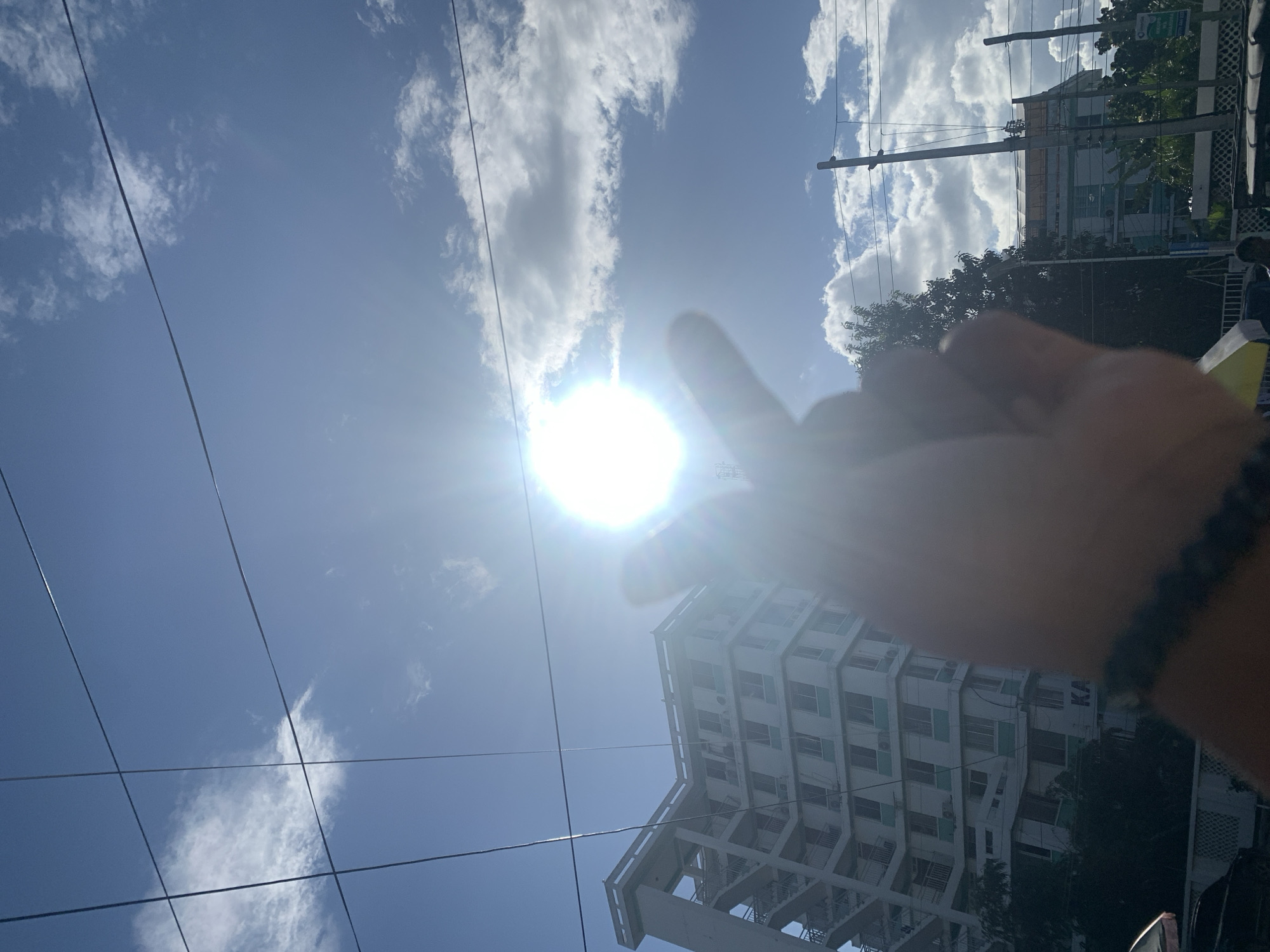 The image, rotated 90 degrees counter-clockwise, displays a bright, sunny outdoor scene under a clear blue sky with scattered white clouds. The central focus is a person's right hand, silhouetted against the intense glare of the sun, which appears as a radiant white orb with prominent lens flare. The hand wears a dark, possibly beaded, bracelet on the wrist, and its fingers (specifically the thumb and index finger) are positioned as if making a gesture, possibly a Korean finger heart, or simply shielding the lens from the direct sunlight.
Overhead, multiple dark utility wires or power lines traverse the sky, connected to a utility pole visible on the right side of the frame. In the background, a modern, multi-story building with a light-colored facade and numerous windows dominates the lower right portion of the image. The building features distinct architectural elements, possibly exterior staircases or balconies. Some dark green trees and foliage are visible near the building and utility pole. Faint text, "KA", can be discerned on the side of the building. The bright sun indicates it is midday or early afternoon. The scene depicts an urban or suburban setting in Dar es-Salaam, Tanzania, under clear and bright weather conditions.