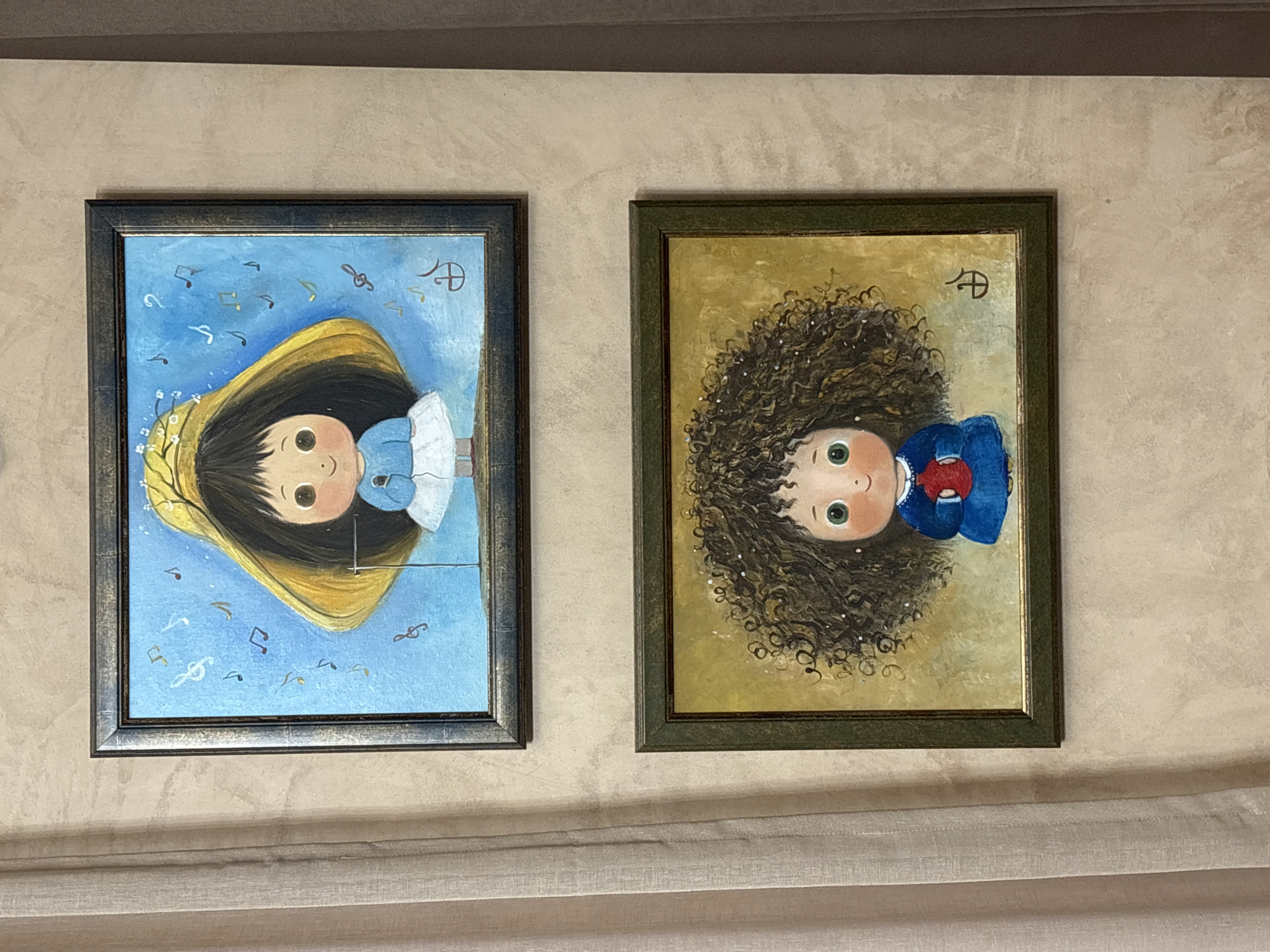 Two framed portrait paintings hang on a beige wall. The painting on the left features a girl with dark hair and a yellow halo, dressed in blue and white, against a blue background with musical notes. The painting on the right shows a girl with curly brown hair, dressed in blue and red, against a golden-brown background. Both paintings have a stylized signature in the upper right corner. The location is possibly a residence or gallery in Tbilisi, Georgia.