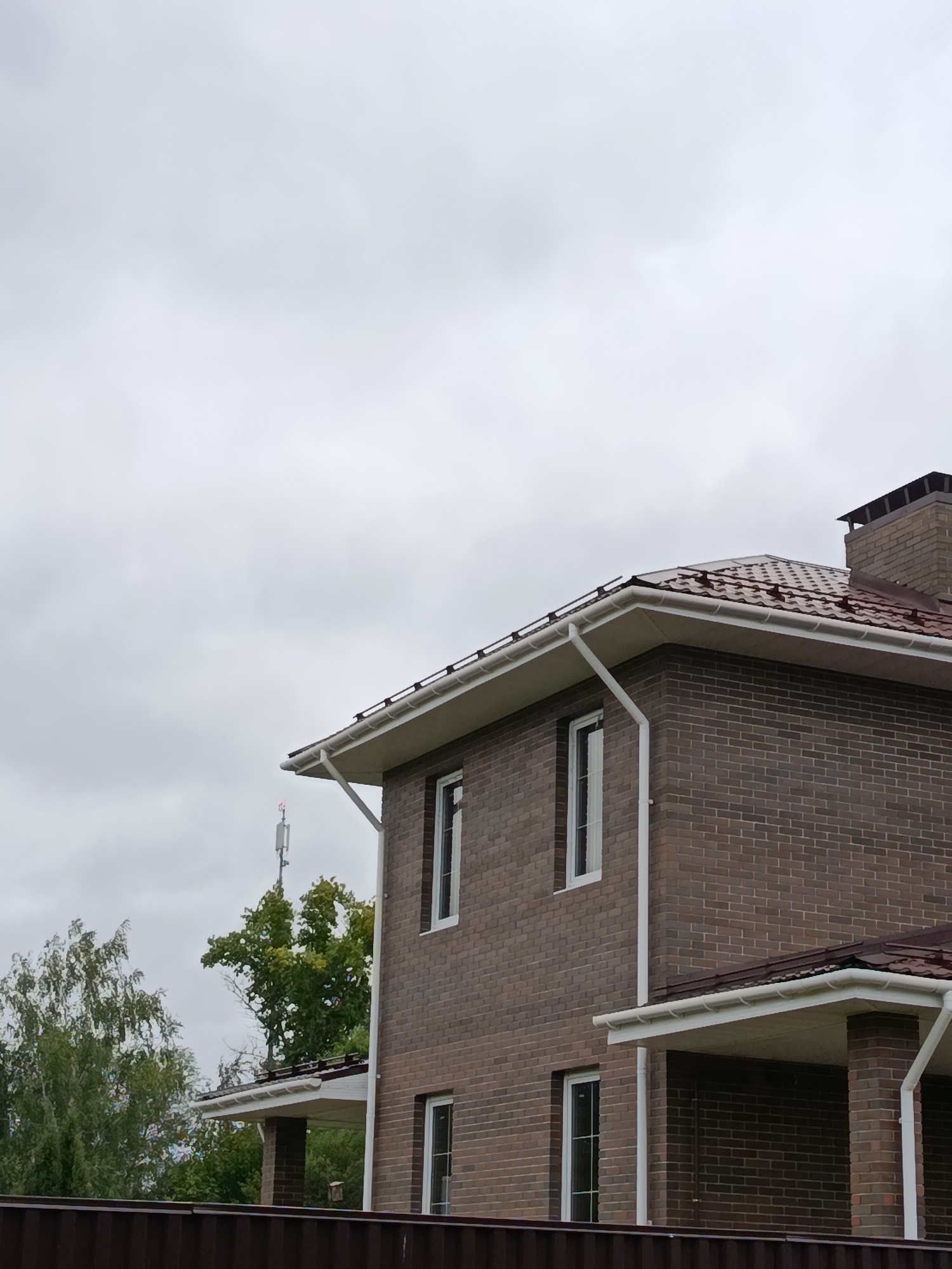 The image shows a two-story brick house with a brown metal roof, white gutters, and white-framed windows, situated in the city of Verëya, Russia. The house is viewed from a low angle, with the sky taking up the upper portion of the frame. To the left of the house, there are trees with green foliage, and in the background, a cell tower with red lights is visible. In the foreground, a dark brown fence runs horizontally across the bottom of the image. The sky is overcast with gray clouds. There are no people or animals visible in the scene.