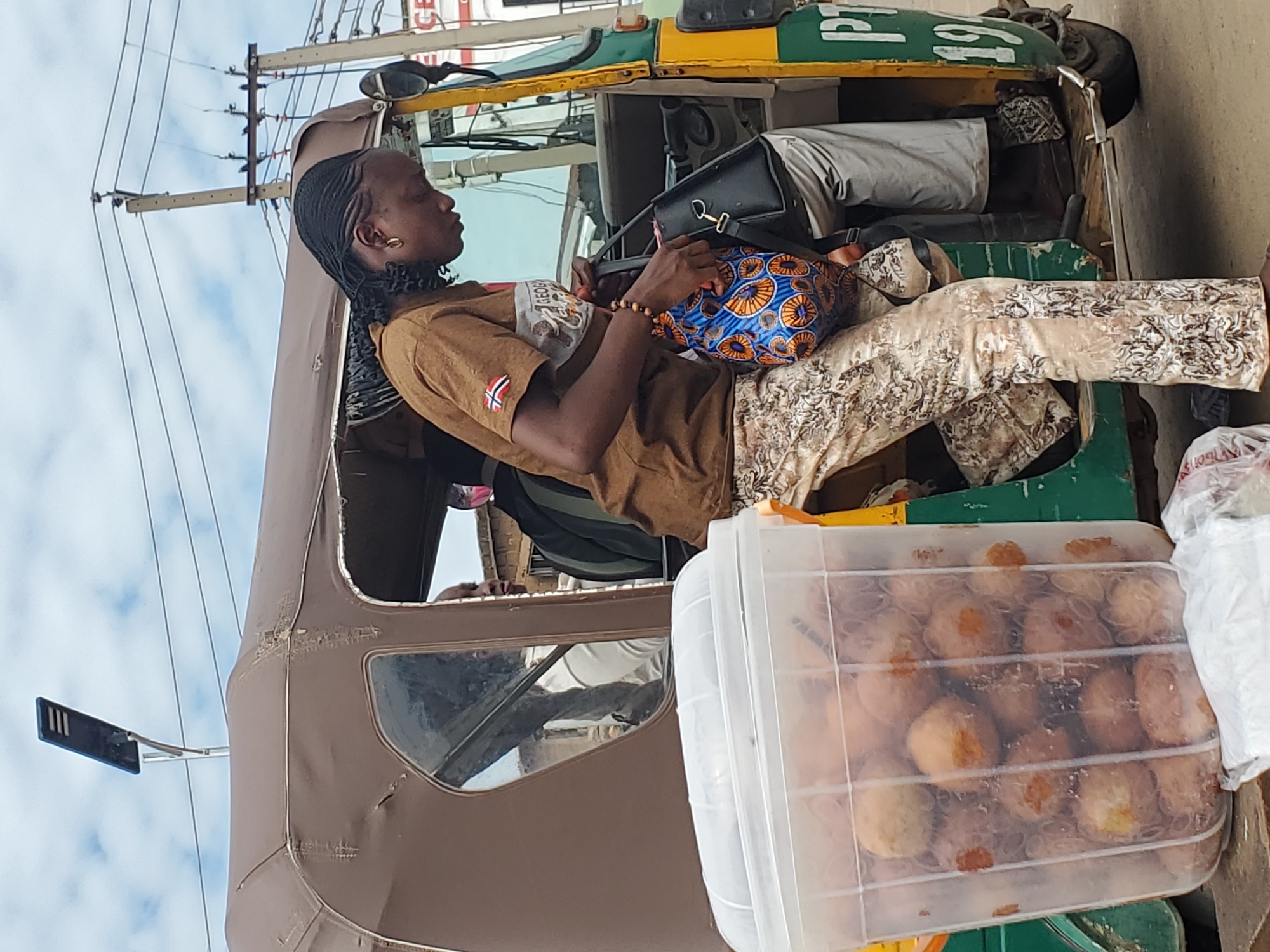 In Gwong, Nigeria, a woman with braided hair is seen disembarking from a brown and green auto-rickshaw. She is wearing a brown t-shirt and patterned trousers, and is holding a black handbag and a colorful patterned tote bag. A large, clear plastic container filled with round, fried food items is positioned in the foreground to the left of the auto-rickshaw. The background shows a street scene with power lines and a solar-powered lamp post.