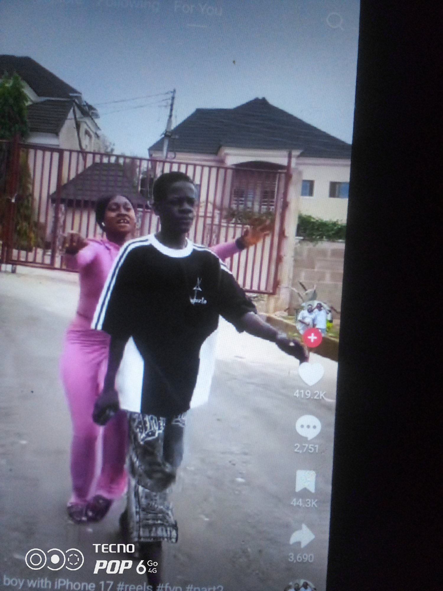 In a street scene, two young people are captured in motion. A female, dressed in a vibrant pink tracksuit, extends her left arm forward, her mouth open in what appears to be a vocalization or exclamation. Beside her, a male figure, wearing a black t-shirt with white stripes and patterned pants, is also in motion, his left arm extended. Behind them, a building with a dark, tiled roof and a brown metal fence can be seen. The ground is paved with what appears to be asphalt or concrete. To the right of the main subjects, a small circular image displays two individuals in white clothing. Social media interaction icons are visible on the right side of the screen, indicating engagement metrics such as likes (419.2K), comments (2,751), shares (44.3K), and plays (3,690). A watermark at the bottom reads "TECNO POP 6 4G" along with text that appears to be related to a video title or description, including "#reels" and "#fyp". The context of the image suggests it was taken in Howayi, Nigeria, within an urban environment.