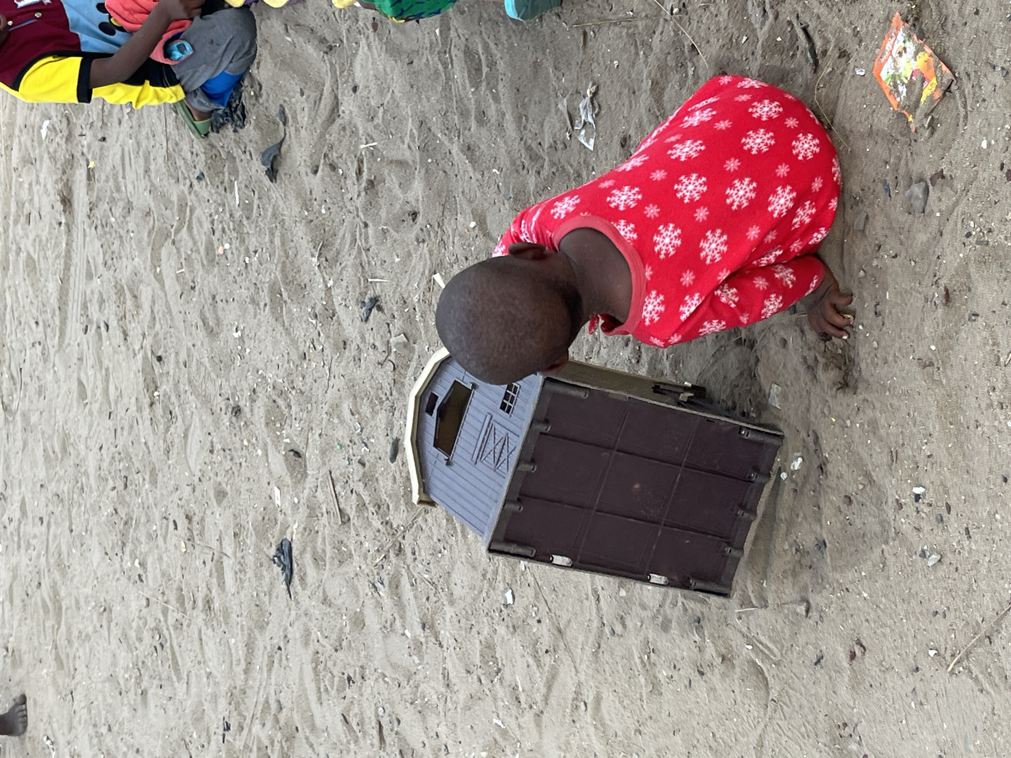 The scene depicts a child in red pajamas with white snowflake patterns kneeling on a sandy surface, likely a beach, next to a brown and grey toy barn. The child appears to be examining the barn. Another individual, partially visible, is wearing a yellow and black sleeve and grey pants. The sandy ground is littered with small debris and remnants of plastic. The location is Badagry, Nigeria.