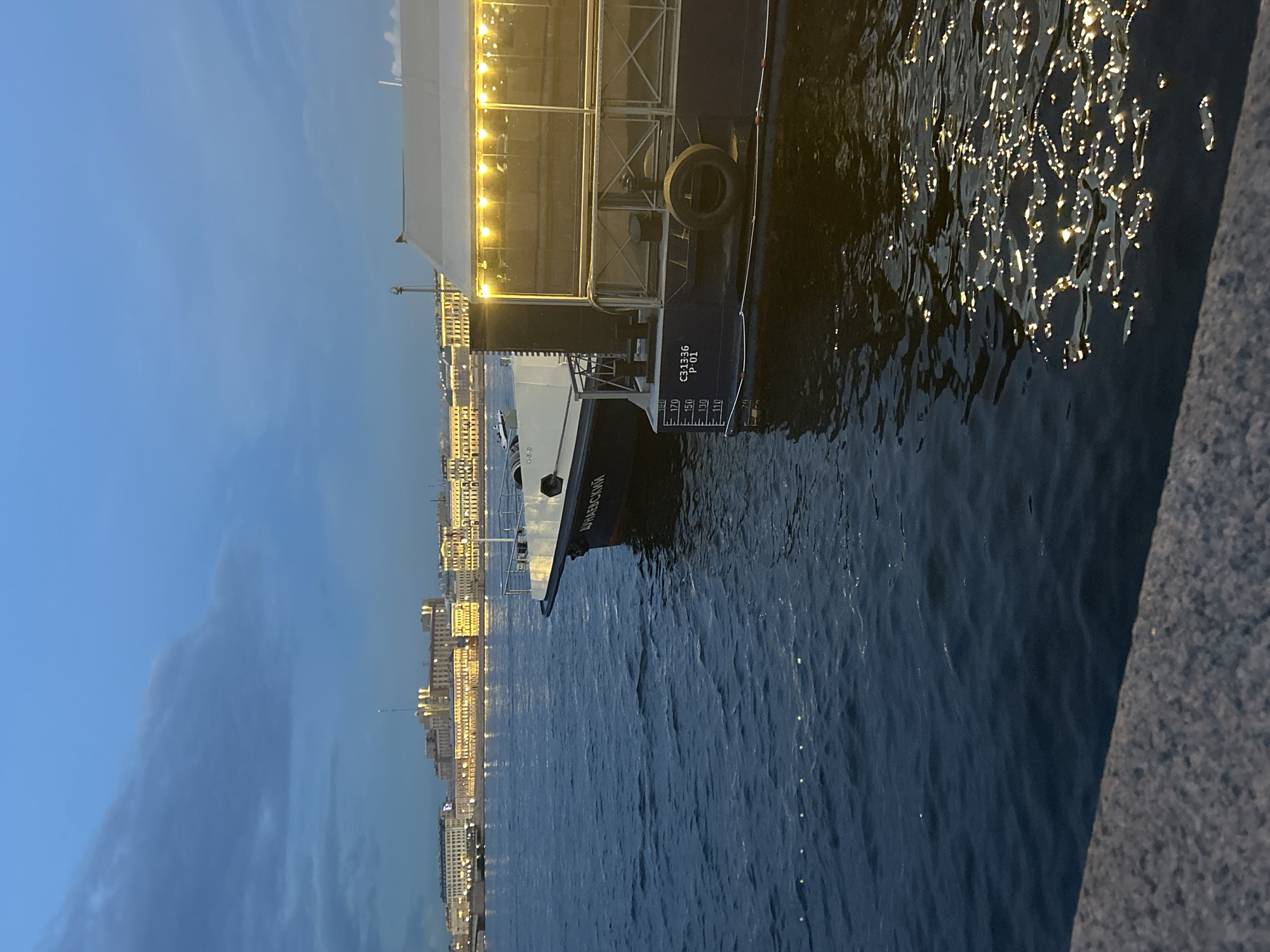 The image depicts a dark, atmospheric scene on a body of water, likely a river or canal, at dusk or early night. In the foreground, the dark, rippling water is illuminated by the reflection of lights from a boat and the distant city skyline. A concrete embankment or pier forms the bottom edge of the frame.
The primary subject is a dark-hulled boat docked or moored. The boat has a white upper deck with a transparent enclosure, lit by a string of warm, yellow lights. The side of the boat is visible, showing dark blue paint and some markings, including depth indicators numbered 70, 110, 130, 150, and 170. There are also numerical and alphabetical markings that read "C31336 P 01". A spare tire is mounted on the side of the hull. The bow of the boat is pointing towards the right of the frame.
In the background, across the water, a city skyline is visible. The buildings are lit with many yellow lights, suggesting a populated urban area. The sky above is a deep blue, with some wispy, darker clouds contributing to the twilight ambiance. The overall mood is serene and slightly moody. The location is identified as Unknown, Unknown.