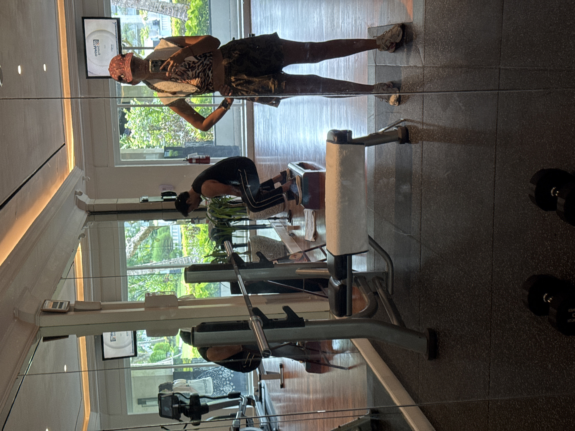 The image shows a person taking a selfie in a gym. The gym has weight machines, windows, and dumbbells on the floor. A person is using a weight machine. The location is Kuta Selatan, Indonesia.