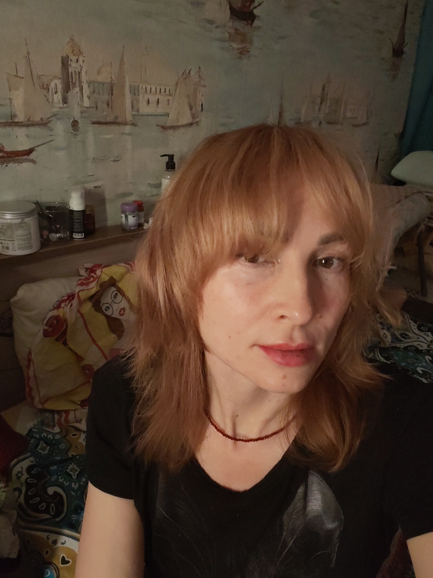 A selfie of a woman with light brown hair and bangs, wearing a black t-shirt and a red beaded necklace. She is indoors, seemingly in a bedroom or personal space. Behind her is a shelf with various bottles and jars. A bed with patterned bedding and pillows is also visible. The wallpaper features a nautical scene with boats and buildings, reminiscent of Venice. The location in the city of Chaikovsky, Russia is not evident from the image.
