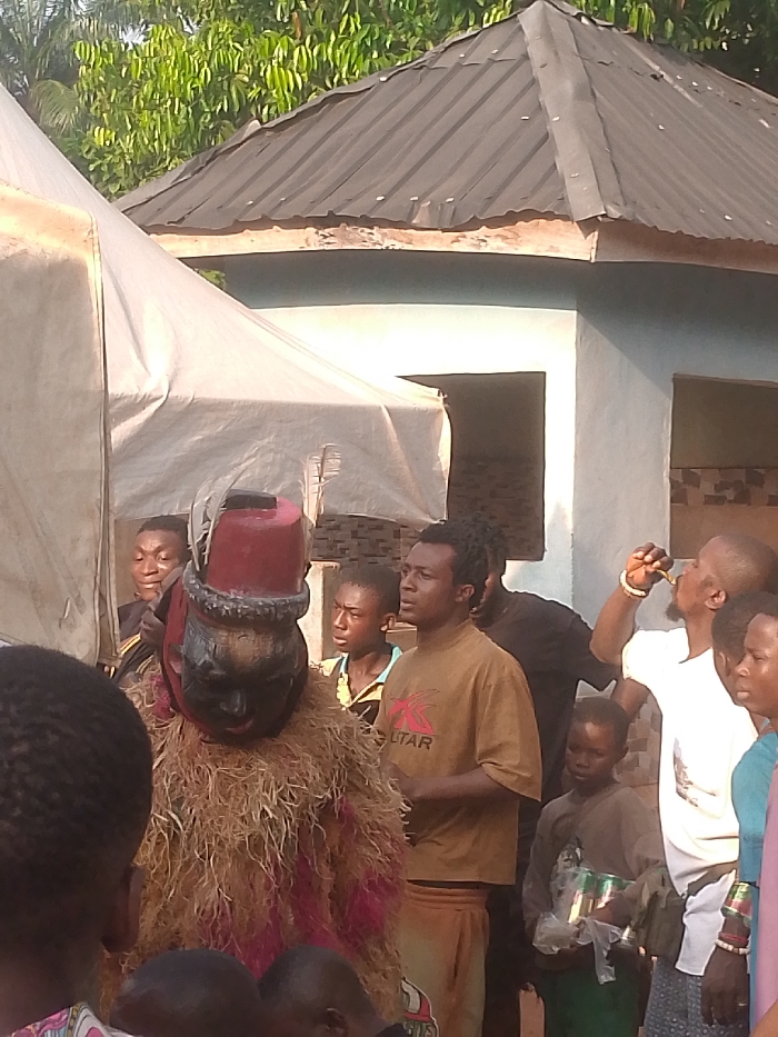 An outdoor scene in Nnobi, Nigeria, depicts a gathering around a central masquerade figure. The masquerade, located left-of-center, wears a dark, carved mask, a red cylindrical headdress with a white plume, and a shaggy, light brown body costume with red accents. Surrounding the masquerade are several individuals of varying ages. A young man with braided hair, wearing a light brown t-shirt with a "KUTAR" logo, stands to the masquerade's right, looking upwards. Further right, a man in a white t-shirt and gold wristband drinks from a small, dark bottle. Another child in a dark shirt and green trousers holds a plastic bag containing multiple metallic cans. Several other people, some partially obscured, observe the scene. In the background, a white canvas tent stands left-of-center, partially obscuring a light blue building with a dark, corrugated metal octagonal roof. Lush green foliage is visible behind the building under bright daylight.