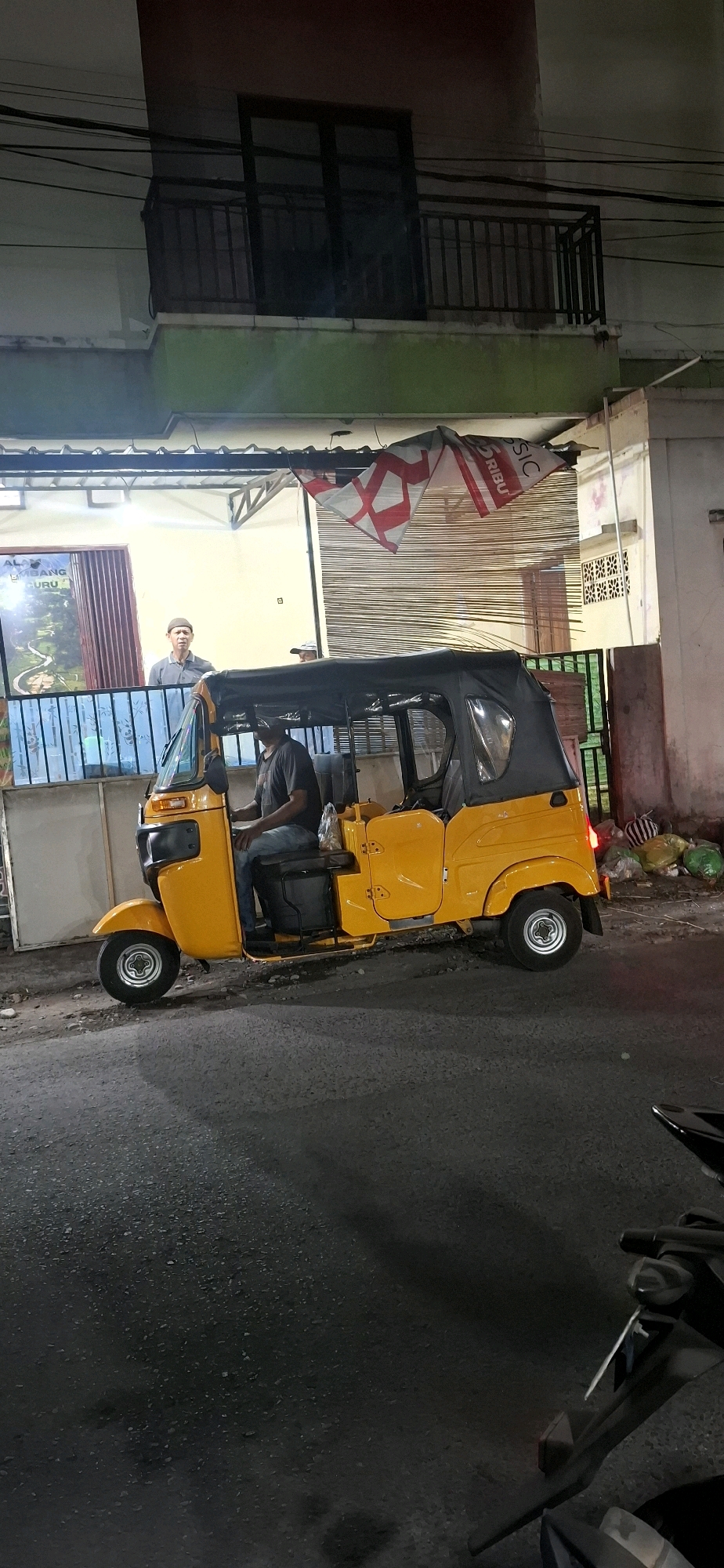 A yellow auto-rickshaw, also known as a becak motor, is parked on an asphalt road in front of a building at night. A man wearing a dark t-shirt and jeans is seated in the driver's position of the auto-rickshaw. Another man, wearing a grey t-shirt and a prayer cap, is standing behind a counter to the left of the auto-rickshaw, looking towards it. The building has a green-painted lower section and a balcony with a railing on the upper level. A banner with red and white diagonal stripes and the word "SIC" is hanging from the building's facade. Trash bags are piled near the building to the right of the auto-rickshaw. The scene appears to be on a street in Surakarta, Indonesia, given the presence of the auto-rickshaw and the architectural style.