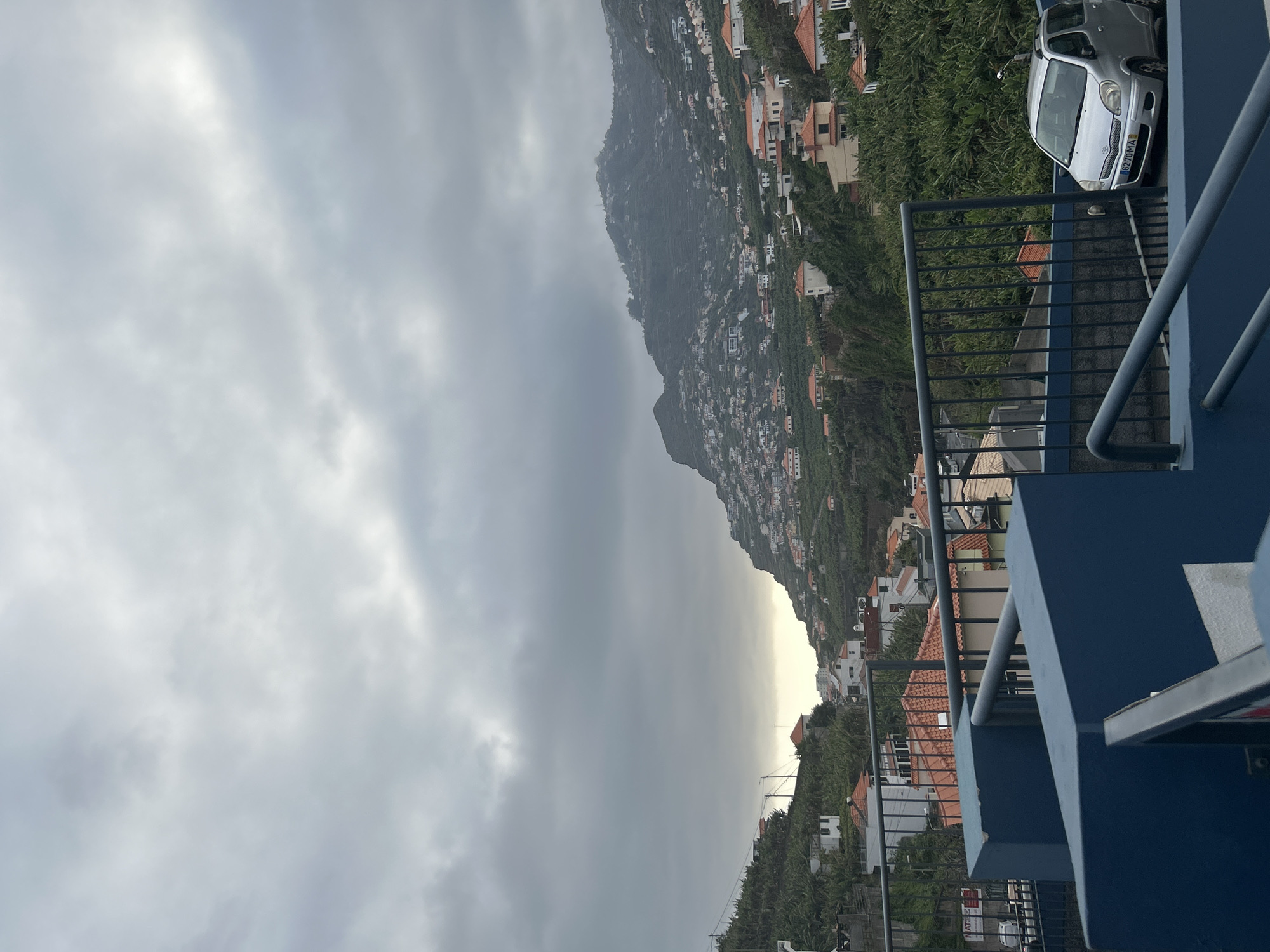 The image captures a view from a balcony overlooking the city of Câmara de Lobos, Portugal, on a cloudy day. The scene features a hillside densely populated with houses and vegetation. A silver car is parked on a platform near the balcony. The sky is overcast, with a section of brightness visible on the horizon. Balcony railings and structural elements are in the foreground.