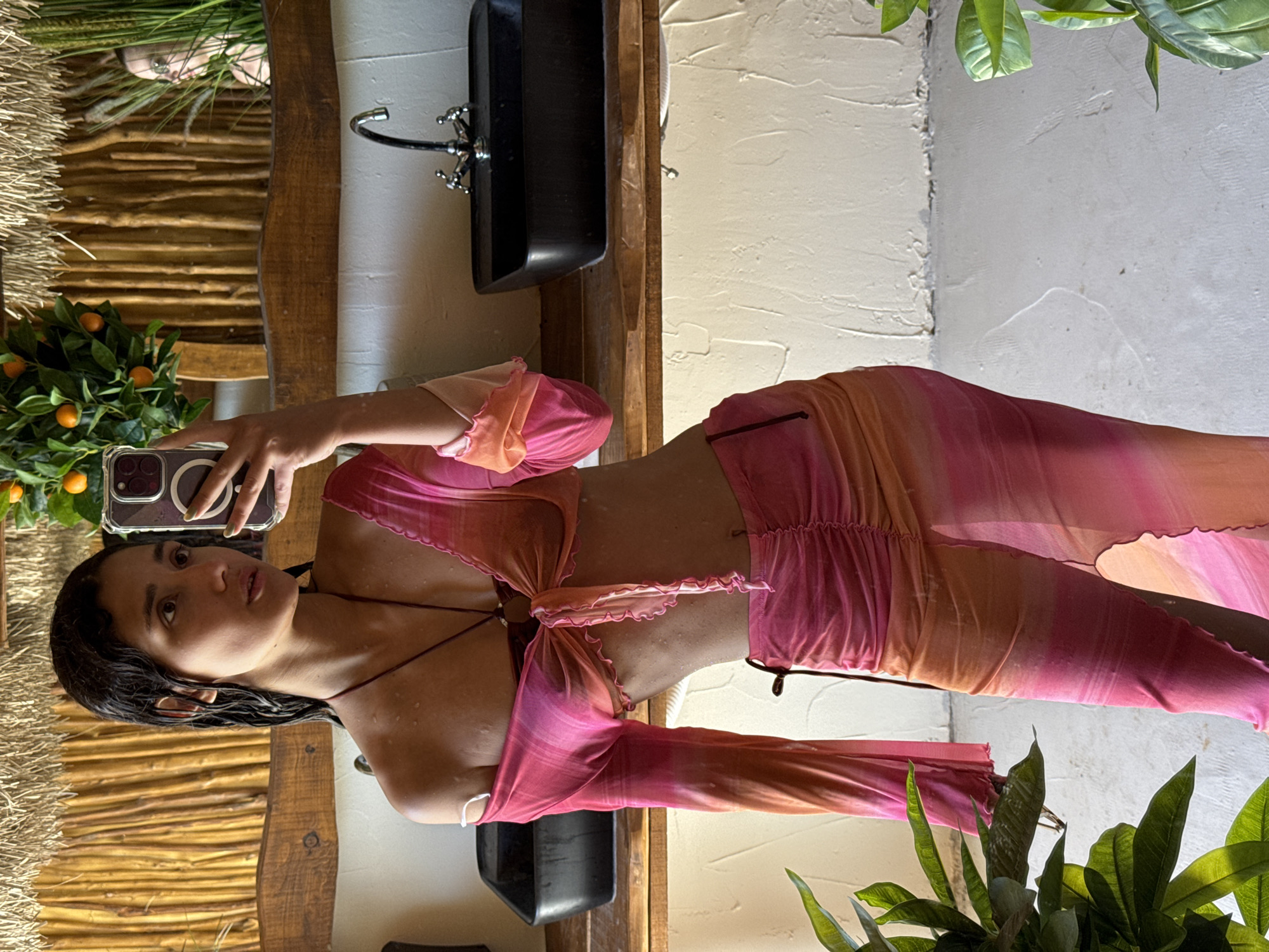 The scene depicts a female individual taking a mirror selfie in an interior setting located in Vrba, Montenegro. The subject, positioned centrally, has dark, wet hair pulled back, with visible water droplets on her skin, particularly on her shoulders and chest. She is wearing a sheer, tie-dye or ombre two-piece outfit consisting of a long-sleeved crop top, tied at the front to expose her midriff, and a matching ruched, high-slit maxi skirt, both in shades of pink and orange. Her left hand, holding a smartphone with a clear case, is extended to capture the reflection. She is wearing a thin, dark necklace.
The immediate background, reflected in the mirror, features a rustic wooden wall composed of vertical slats or branches, topped with a thatched or woven trim. A small, artificial plant bearing orange fruit, resembling tangerines, sits on a ledge to the left of the frame, reflected near the subject's head. To the right, another green, leafy plant with long blades is reflected, and a faint, blurry reflection of a human figure is visible behind it.
In the foreground, a rough-hewn wooden countertop supports two rectangular, black, above-counter sinks. A chrome faucet with two cross-handle taps is installed at the rear of the right sink. The wall below the counter is textured and off-white. Green leafy plants partially obscure the lower left and right corners of the frame. The floor appears to be a light-colored, textured surface.