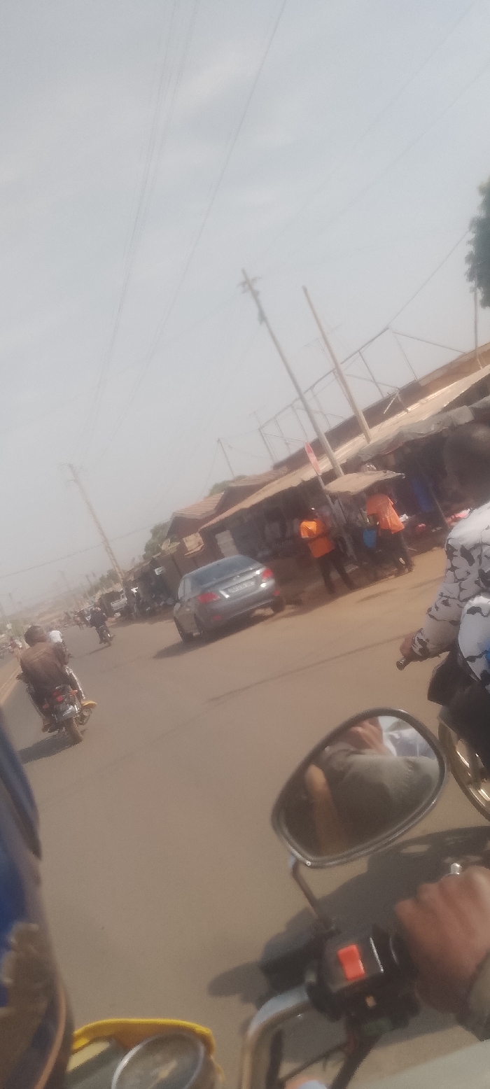 The image depicts a daytime street scene in Korhogo, Côte d'Ivoire, captured from the perspective of a motorcycle rider. In the immediate foreground, elements of a motorcycle are visible, including a blue and yellow section on the left, a speedometer/gauge, the rider's right hand gripping the handlebar, an orange control switch, and a right rearview mirror reflecting the upper body of an individual. The asphalt road stretches ahead, populated by traffic. On the left side of the road, a person in a brown jacket rides a motorcycle, followed by another motorcycle in the distance. On the right side of the road, a silver sedan (license plate "4490 GG 01") is positioned near the curb. Along the right-hand side of the street, multiple roadside stalls with corrugated metal roofs are visible, indicating a commercial area. Several individuals are present near these stalls; at least three are distinctively wearing bright orange shirts. Further in the background, additional structures with reddish-brown roofs are visible. Overhead, multiple utility poles support power lines against a hazy, light grey sky.