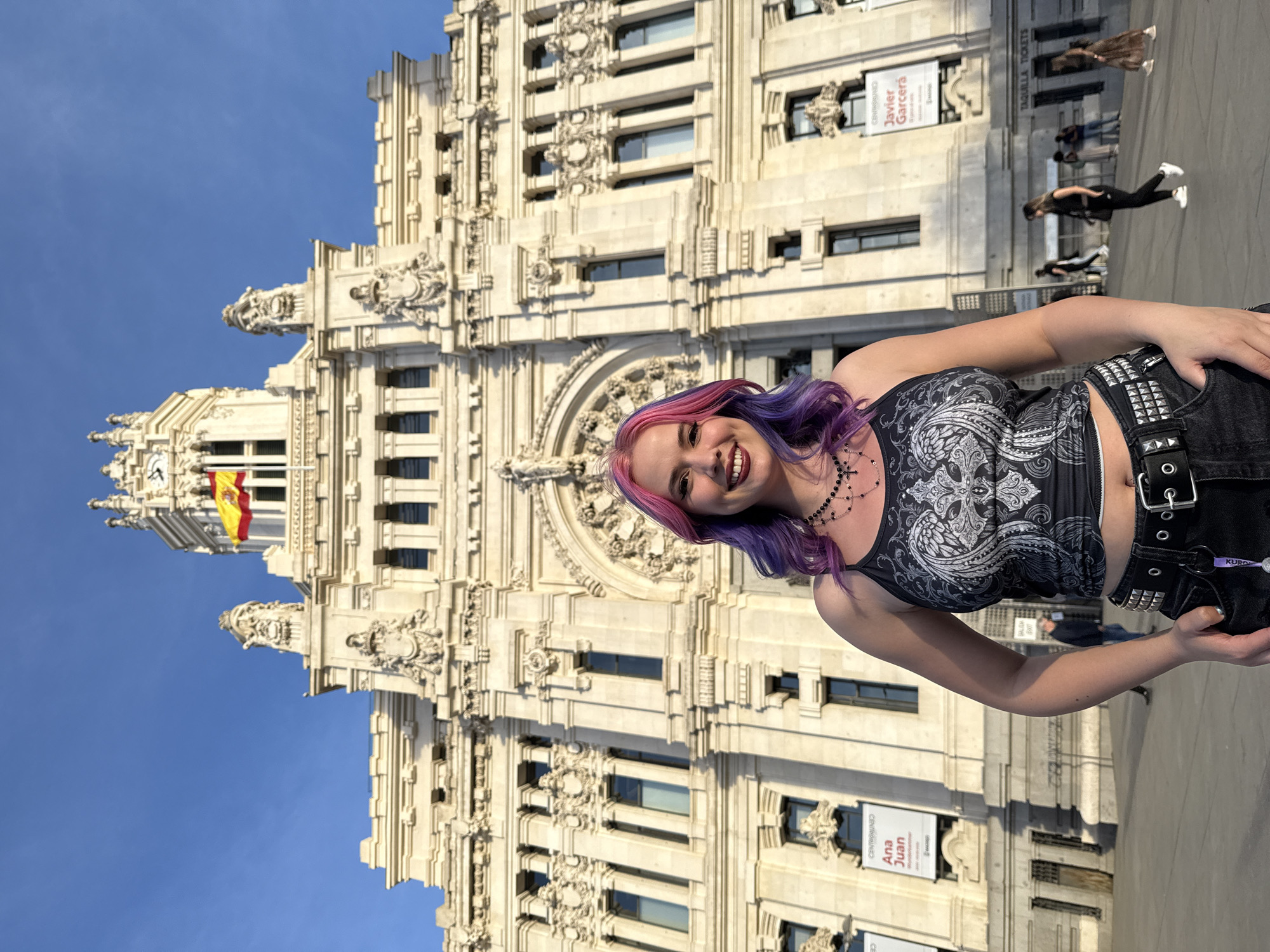 The image depicts a woman with dyed pink and purple hair standing in front of a grand, ornate building. The building, identifiable as the Palacio de Cibeles in Madrid, Spain, has intricate architectural details. The Spanish flag is visible atop the building. The woman is wearing a dark top and jeans with a studded belt. Several signs are attached to the building's facade. Other people can be seen in the background on the pavement around the building. The sky is clear and blue.