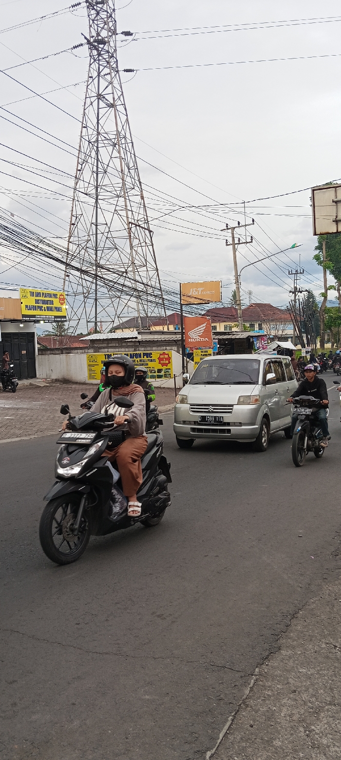 The image shows a street scene in Cangkuang, Indonesia, located at -7.044 latitude and 107.56 longitude. In the foreground, a woman wearing a brown hijab and a black face mask is riding a black motorcycle. The motorcycle is in the left lane of the street. Behind the woman, a white minivan is driving in the right lane. A man on a black motorcycle with a helmet and a green jacket is stopped behind the minivan. In the background, a large metal electrical tower stands tall with numerous power lines spanning across the sky. The road is asphalt and the sidewalk is concrete. The sky is overcast with a few wispy clouds. There are signs for shops and businesses in the background. Several trees and buildings are visible. The woman is riding the motorcycle towards the viewer.