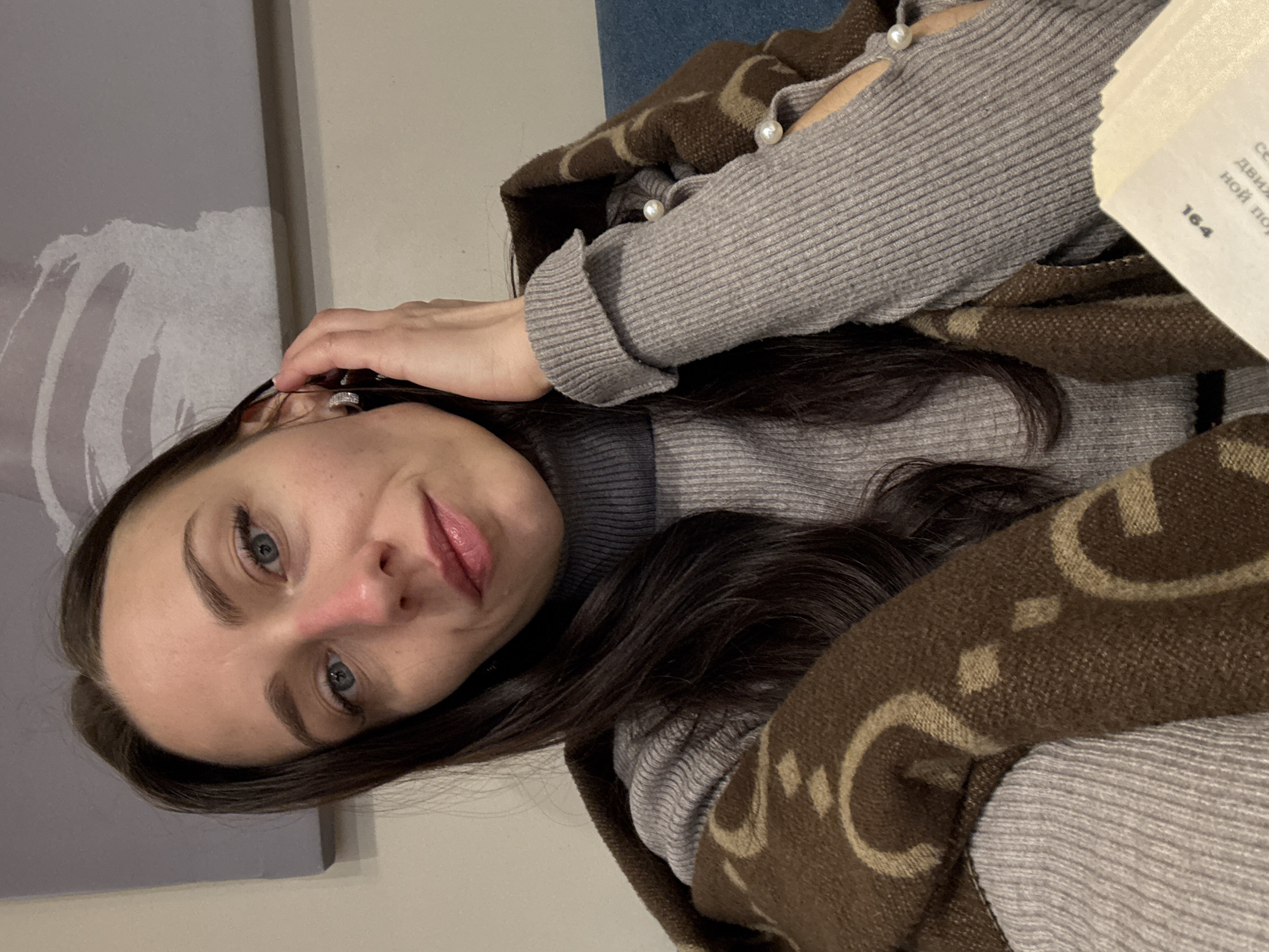 An indoor portrait from Saint Petersburg, Russia, shows a woman with long, dark hair and blue eyes looking directly at the camera with a gentle, calm expression. She is wearing a light gray ribbed long-sleeved top with pearl-like embellishments on shoulder cutouts. A brown blanket or scarf with a light brown pattern is draped around her shoulders and arm. Her right hand is visible near her head.
The background features a light-colored wall and a portion of what appears to be abstract artwork or a painting on the left. A hint of blue fabric or furniture is visible in the upper right corner. The woman holds a book with visible Cyrillic text, including "СЕВ," "движ," and "НОЙ ПО," along with the page number "164." The scene conveys a relaxed and quiet moment.