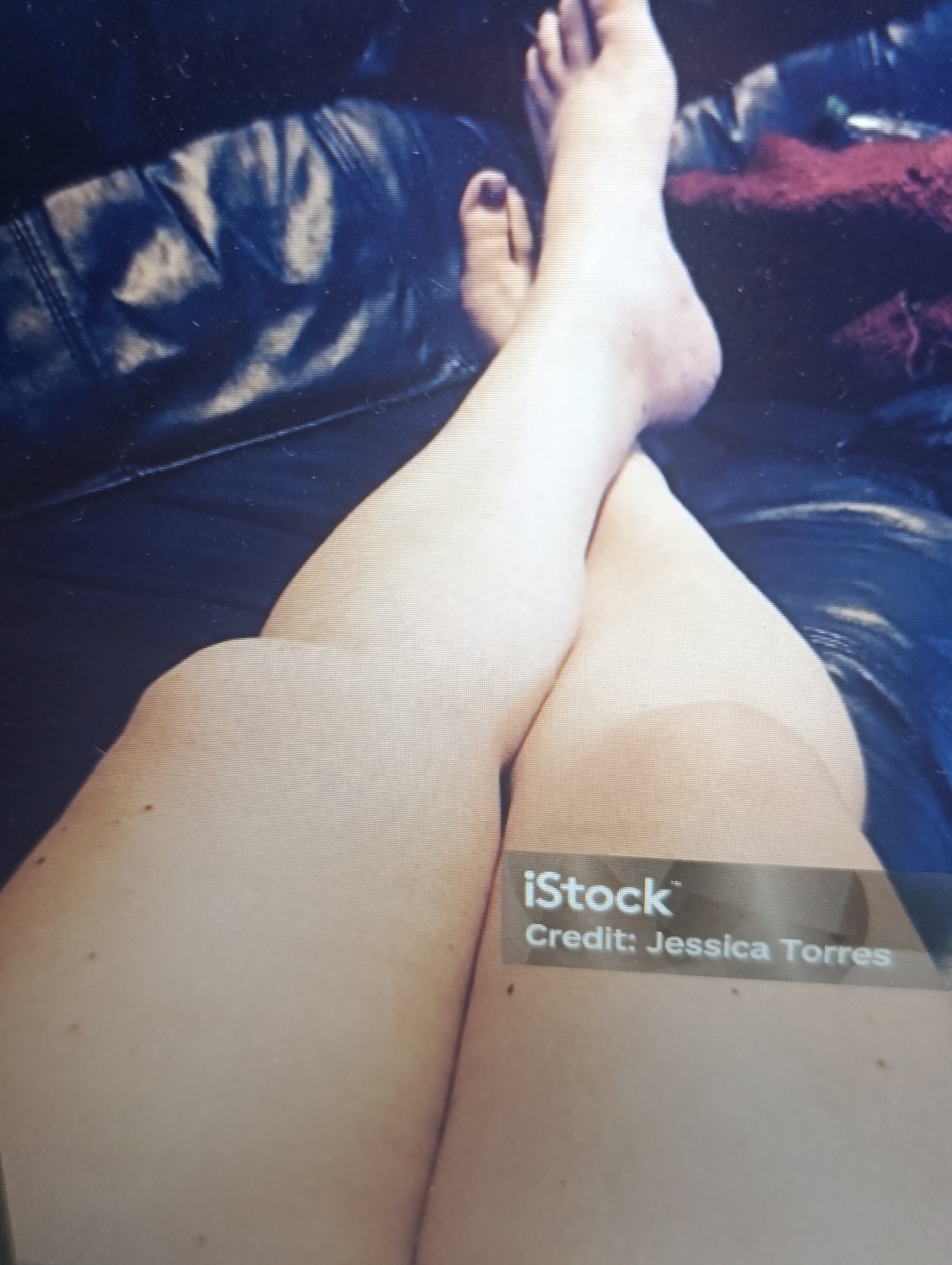 The image shows a close-up of a person's legs, crossed and resting on a dark, possibly leather, surface. The skin tone is light, and there are a few moles visible. The feet are also visible, with one toe nail painted dark red. A red blanket or fabric is partially visible in the background. There is an "iStock" watermark with "Credit: Jessica Torres" overlaid on the lower right portion of the image. The scene appears to be indoors, possibly on a couch or chair, in an unknown location within an unknown city.