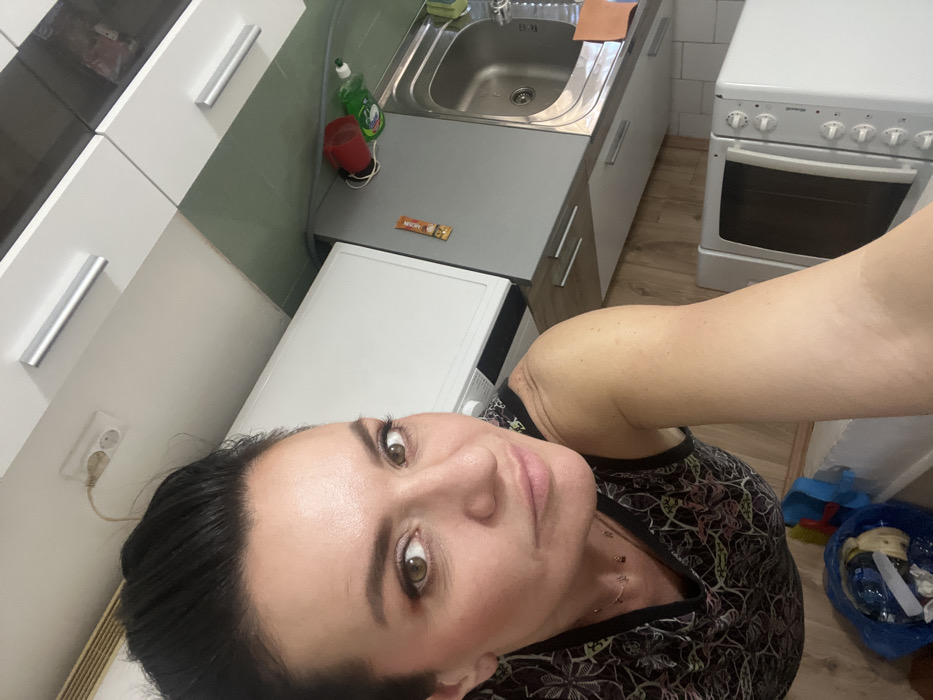 The image displays a rotated indoor scene, approximately 90 degrees counter-clockwise, centered on a woman taking a selfie within a residential kitchen located in Smederevska Palanka Municipality, Serbia.
**Person:**
A woman, appearing to be middle-aged, occupies the left and central portion of the frame. She has dark hair styled away from her face, exposing her forehead and ears. Her skin tone is light, and she wears visible makeup, including eyeshadow, eyeliner, mascara, and blush. Her eyes are brown, and she looks directly at the camera with a subtle expression. She is wearing a dark, sleeveless top featuring a light-colored, intricate abstract or floral pattern. A delicate, gold-colored necklace with small, dark charms is visible around her neck. Her right arm and shoulder are visible in the lower-right quadrant, extending towards the camera, indicating she is holding the device herself.
**Environment and Objects:**
The background reveals a kitchen setting.
* **Upper-left quadrant:** A white double electrical outlet with a light brown power cable plugged into one socket and extending upwards out of frame. This outlet is affixed to a white surface, likely a wall or upper cabinet side.
* **Upper-right quadrant:** White overhead kitchen cabinets with horizontal silver bar handles. One cabinet door appears to be a darker grey or black material.
* **Mid-left:** A white appliance, possibly a washing machine or dishwasher, is partially visible.
* **Mid-right:** A kitchen counter area featuring:
* A dark green wall section, likely tiled or painted, above the counter.
* A grey countertop.
* A built-in stainless steel sink with a faucet.
* Items on the counter near the sink include a red plastic cup, a green bottle (likely dish soap) with a white cap, and a small orange square item (possibly a sponge or cloth).
* A brown rectangular package or bar is attached to the side of a grey base cabinet.
* **Lower-left quadrant:** A blue plastic bag, open at the top, containing assorted refuse, including plastic bottles and other items, rests on the floor.
* **Lower-middle/right:** The floor is covered with light-colored wooden planks.
* **Below the woman's right arm:** A white microwave oven with a dark front door and white control knobs is visible.
The lighting appears to be ambient indoor illumination. No external light sources are directly visible.