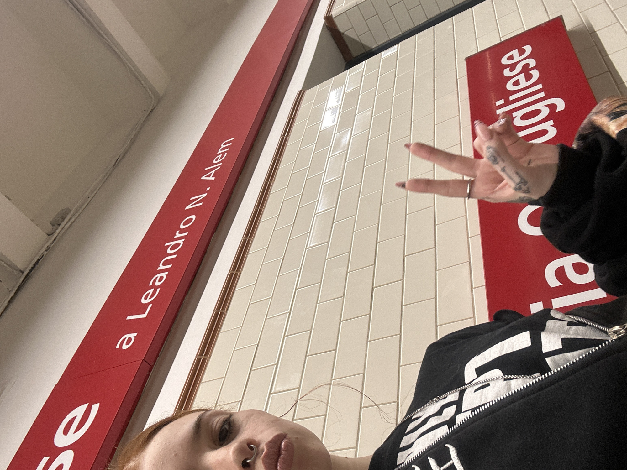 A low-angle, close-up shot captures a person taking a selfie in what appears to be a subway station in Buenos Aires, Argentina. The subject, visible from the chin up, has a nose ring, light-colored hair, and wears a black zip-up hoodie with unidentifiable white text. Their right hand is raised, making a V-sign or peace gesture, revealing tattoos on the fingers and dark-painted nails.
The background features typical subway station elements: a wall covered in light-colored rectangular subway tiles, and prominent red signs with white lettering mounted along the wall. These signs include the partial text "se" on the left, "a Leandro N. Alem" clearly visible on the main sign, and the partial word "ugliese" on a sign to the right, suggesting "Pugliese." The presence of "Leandro N. Alem" strongly indicates the eponymous subway station, a terminus of Line B of the Buenos Aires Subte. The overall setting, with its tiled walls and specific signage style, is consistent with the Buenos Aires metropolitan train system. The time of day and weather are indeterminate as the setting is indoors.