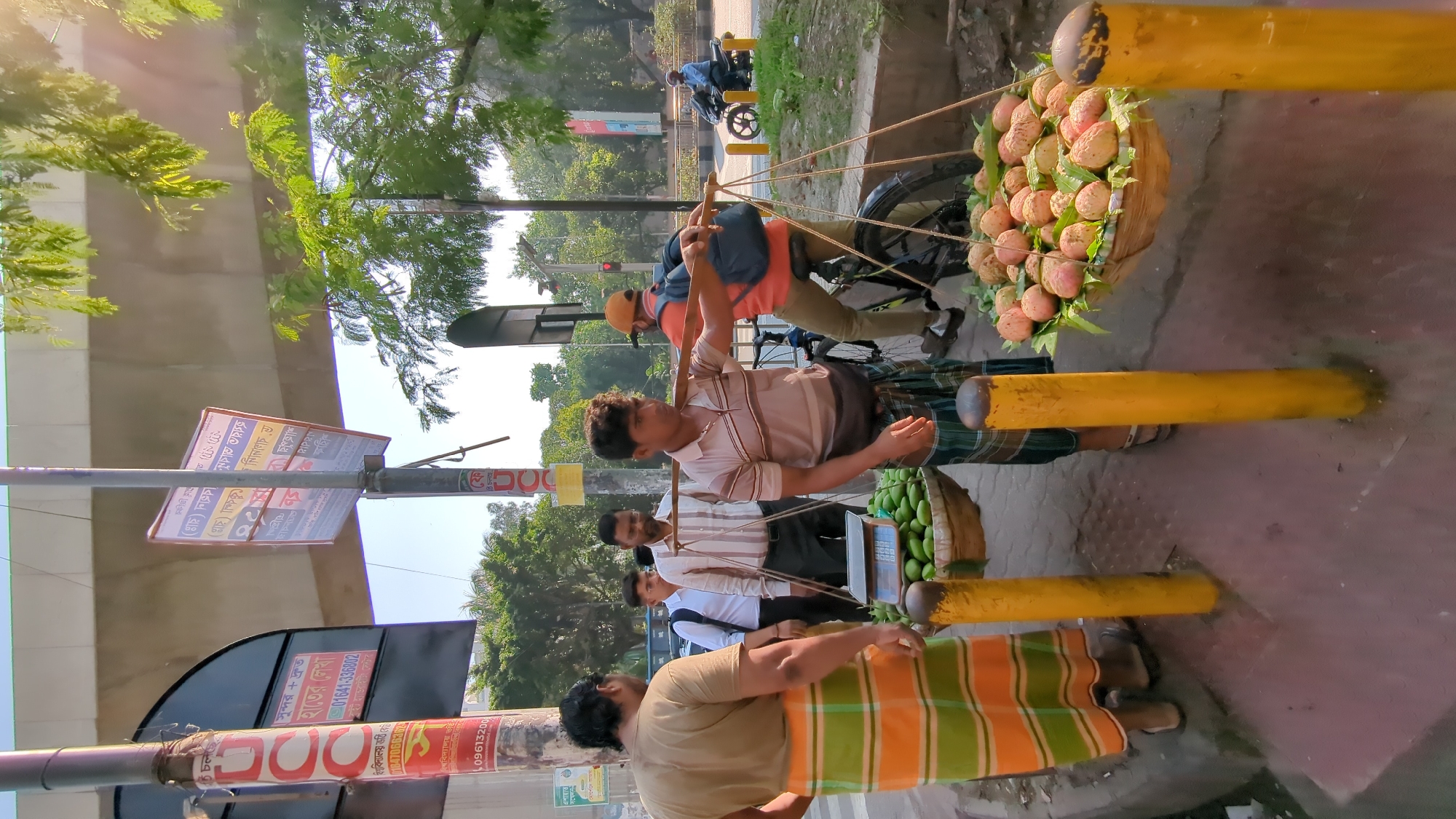 This image captures a street scene in Dhaka, Bangladesh. Several men are present, some wearing traditional lungis. One man is carrying a yoke laden with baskets of fruit, specifically lychees and what appear to be green mangoes. He seems to be a vendor, as does a man in an orange and green lungi. A scale is visible, indicating a commercial transaction. Other individuals are nearby, possibly customers. In the background, one can see a parked motorcycle, trees, and buildings. Signage is visible on poles, likely with local advertisements or information. The overall environment suggests a typical day in a bustling urban area of Dhaka.