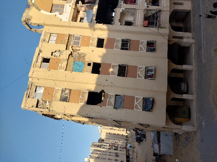 The image depicts a severely damaged multi-story residential building in Khan Yunis, Palestinian Territory, under a clear blue sky. The building, constructed with a light brown facade and reddish-brown horizontal bands, features extensive structural damage, including large perforations, exposed interiors, and shattered or missing windows, some covered with blue tarps or dark material. Ornate architectural details are visible but degraded. On the right side, a balcony on an upper floor has clothes hanging. The ground floor consists of an open arched space, beneath which a green industrial waste bin and a silver sedan are parked. To the left of the main building, the ground is a debris-strewn area with several makeshift shelters constructed from tarpaulins and corrugated metal, and additional damaged multi-story buildings are visible in the background. In the lower right foreground, on a paved surface, two individuals, appearing as men or older boys, stand facing left. Faint figures, possibly children, are discernible in some lower-level windows of the main building. Thin utility wires crisscross the sky. The perspective is slightly elevated.