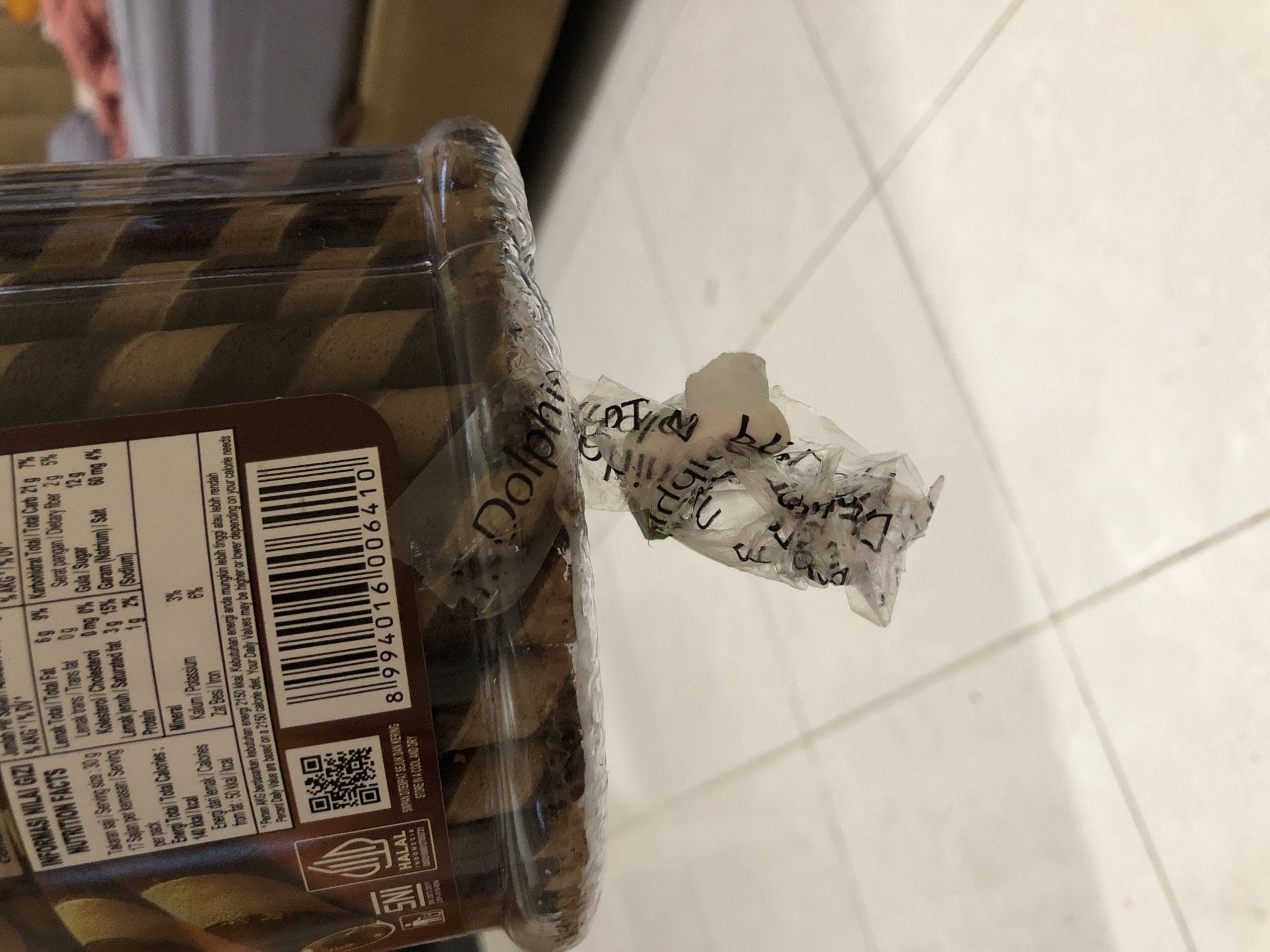 The image shows a close-up of a clear plastic container holding chocolate wafer rolls. The container has a nutritional information label in Indonesian and English. Attached to the outside of the container is a crumpled piece of clear plastic packaging with text and symbols visible. The container is placed on a light-colored tiled surface. The background is blurred and shows what appears to be a bed with a pink blanket. The scene is indoors and does not provide any information about the city of Pashiebeutong, Indonesia.