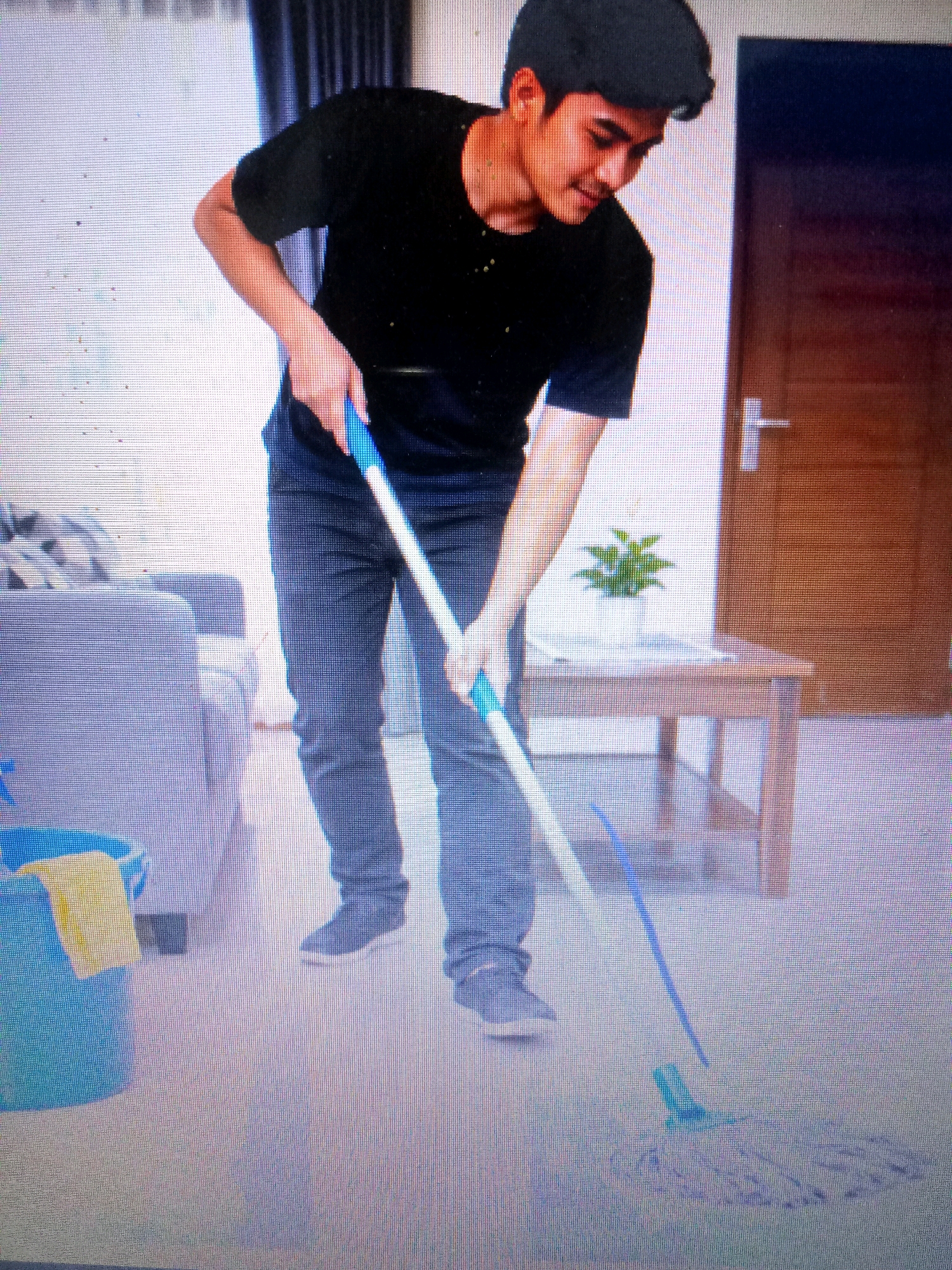 In an indoor residential setting, a young man with dark hair and a black t-shirt is mopping the floor. He is wearing dark jeans and grey sneakers. He is hunched over, actively using a mop with a blue handle and white and blue mop head. To the left of the man, a blue bucket with a yellow cloth is visible. Behind him, a wooden coffee table with a potted plant sits between a gray couch on the left and a wooden door on the right. The floor appears to be light-colored tile or linoleum. The overall environment suggests a clean, modern home. The image does not provide any specific information to identify the location as Bekasi, Indonesia.