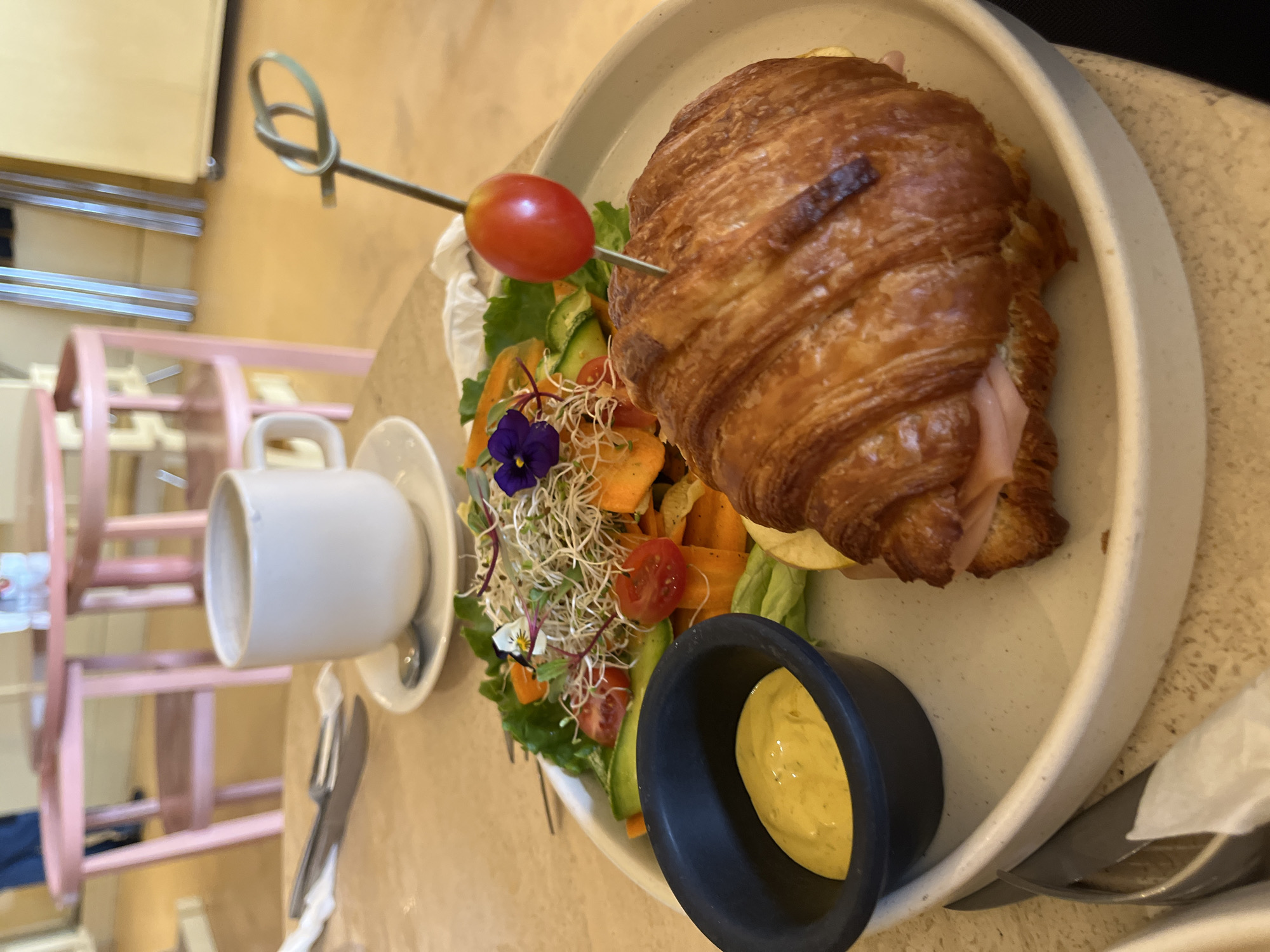 This image depicts a prepared meal set on a table in an indoor dining environment, specifically located in San Andrés Cholula, Mexico. The central subject is a light-colored, circular ceramic plate holding a large, golden-brown croissant sandwich filled with pink ham slices. A thin, dark-colored skewer pierces the croissant, topped with a single red cherry tomato and a green decorative loop. Alongside the croissant is a fresh side salad comprising green leafy lettuce, thinly sliced orange carrots, green cucumber slices, halved red cherry tomatoes, white sprouts, and two small edible flowers, one purple and one with white and yellow petals. A small, matte black ramekin to the left of the salad contains a light yellow, creamy sauce. In the upper left background, a white ceramic mug with a handle rests on a matching saucer, which also holds a silver spoon. To the left of the mug, a silver fork and knife are partially visible, wrapped in a white napkin. The background is blurred, revealing pink wooden chairs and tables, a warm-toned floor, and indistinct metallic structures, indicative of a cafe or restaurant setting. No individuals are visible in the frame.