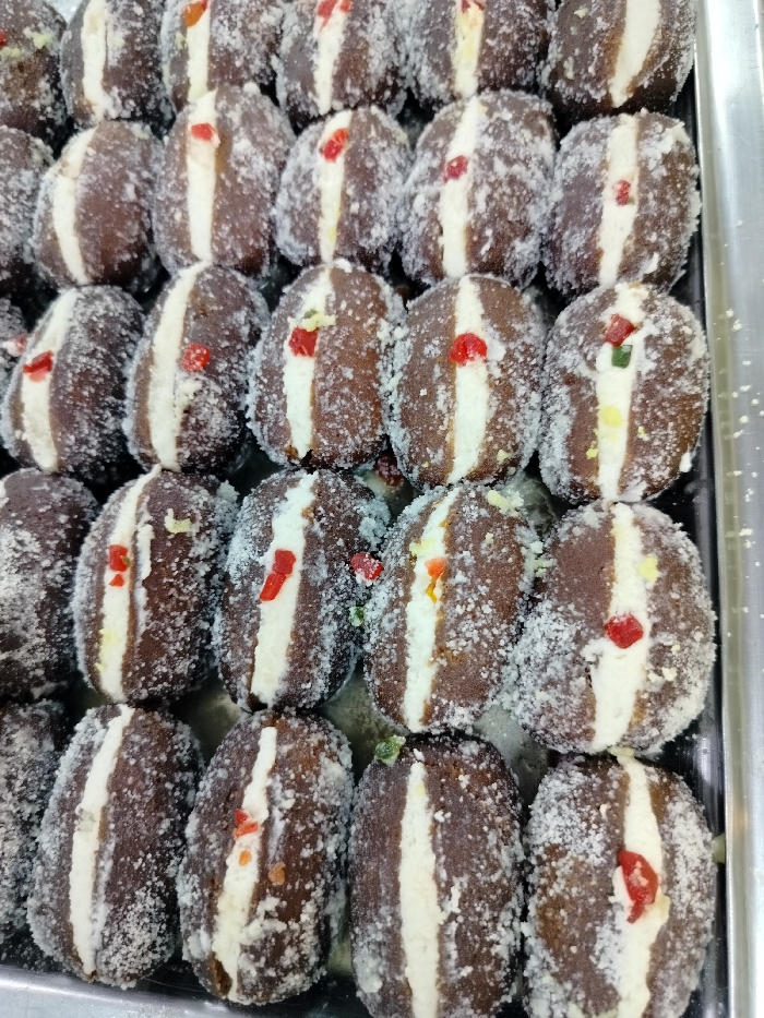 The image shows a close-up view of a tray of Gulab Jamun, a popular Bengali sweet, in Dhaka, Bangladesh. The sweets are arranged in rows and are covered in white sugar crystals. They are dark brown in color with a white filling and are garnished with red, green, and yellow candied fruit pieces. Each Gulab Jamun appears to be approximately 3 cm in diameter and 5 cm in length. They are displayed on a silver tray.