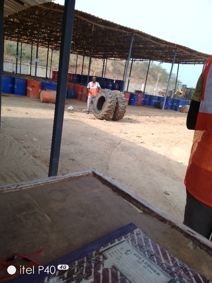 The scene depicts an outdoor industrial storage area in Hoche, Nigeria, under bright daylight. In the mid-ground, an adult male, wearing a white shirt, light trousers, and an orange high-visibility vest, stands near two large, heavily-treaded vehicle tires leaning against each other on dusty earth. Behind him, under a large, open-sided canopy structure with a metal framework, numerous industrial drums are stacked in rows; these include blue drums, some marked 'SERVO,' and orange/red drums. A yellow piece of heavy machinery, possibly a front-end loader, is visible in the far background. In the immediate foreground, a partial view of a weathered rectangular surface, consistent with a desk or counter, displays a ledger book labeled "600 LEDGER" and red wired headphones. A dark, vertical metal support beam occupies the left foreground. The environment consists of dry, light-colored soil and distant sparse vegetation on a hillside.