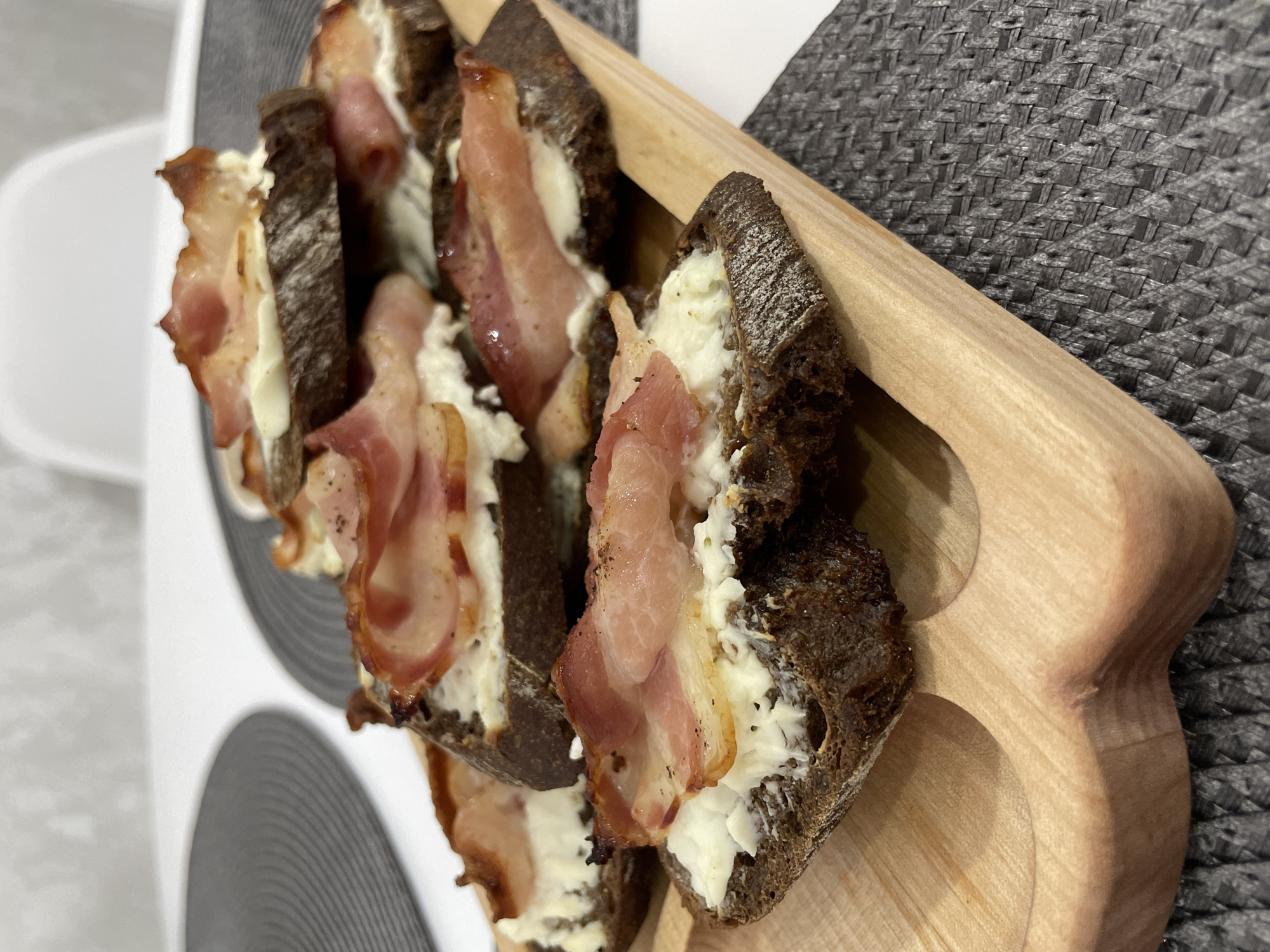 The image shows several slices of dark bread topped with a white creamy substance and bacon strips, arranged on a wooden serving board. The board rests on a surface with a woven gray pattern. Another surface, patterned with concentric gray and white circles, is visible in the background. The location and city of Izhevsk, Russia cannot be determined from the image. There are no people or interactions visible. The environment consists of surfaces and food items.