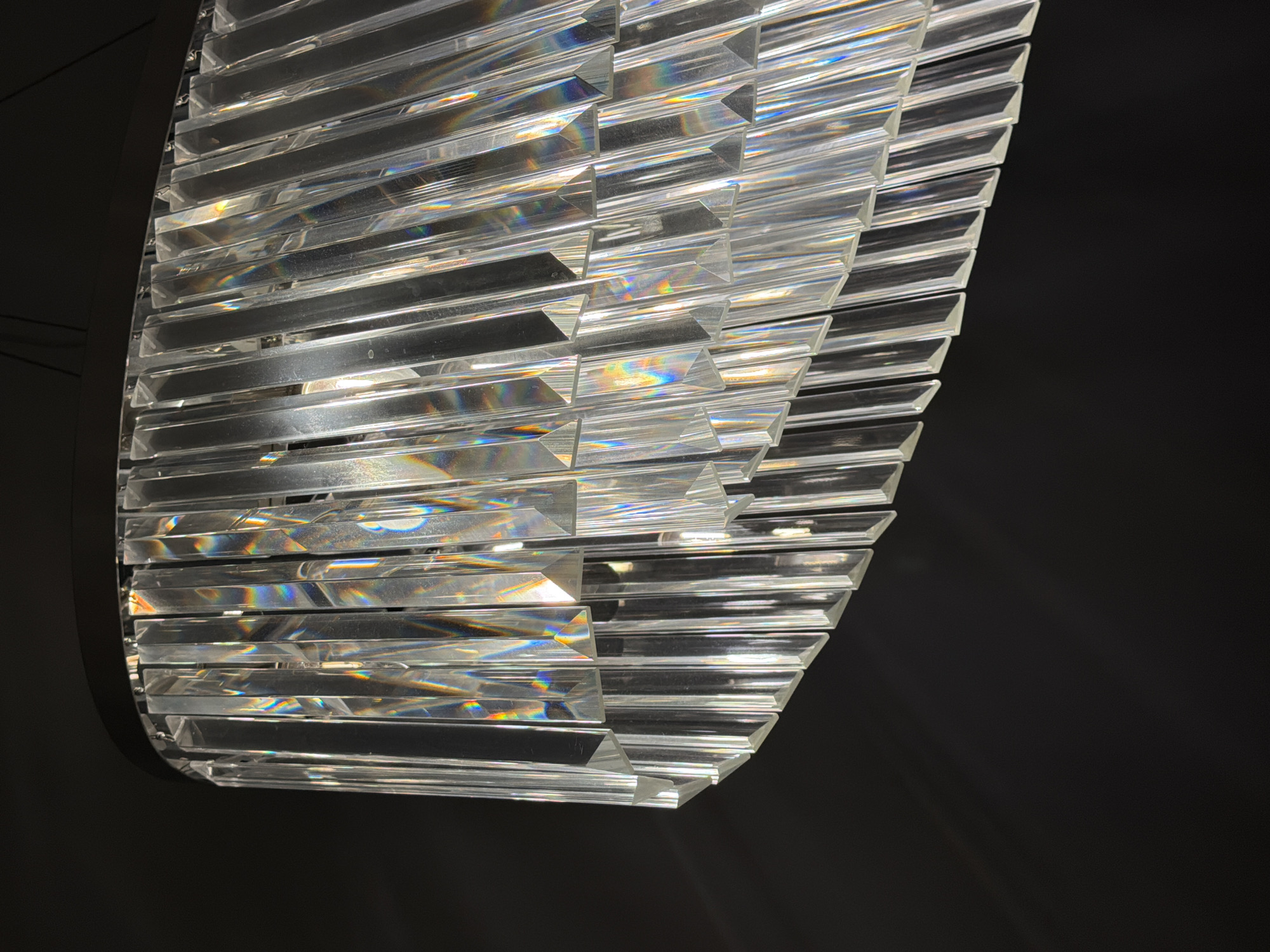A close-up shot depicts a modern chandelier with horizontal, rectangular prisms arranged in a cascading, layered pattern. The prisms appear to be made of clear acrylic or glass. The light reflecting and refracting through the prisms create iridescent patterns of color. The background is a dark, solid color, likely black, which provides a stark contrast and highlights the form and details of the chandelier. There is no evidence or indication to support the image was taken in Romashkovo, Russia.