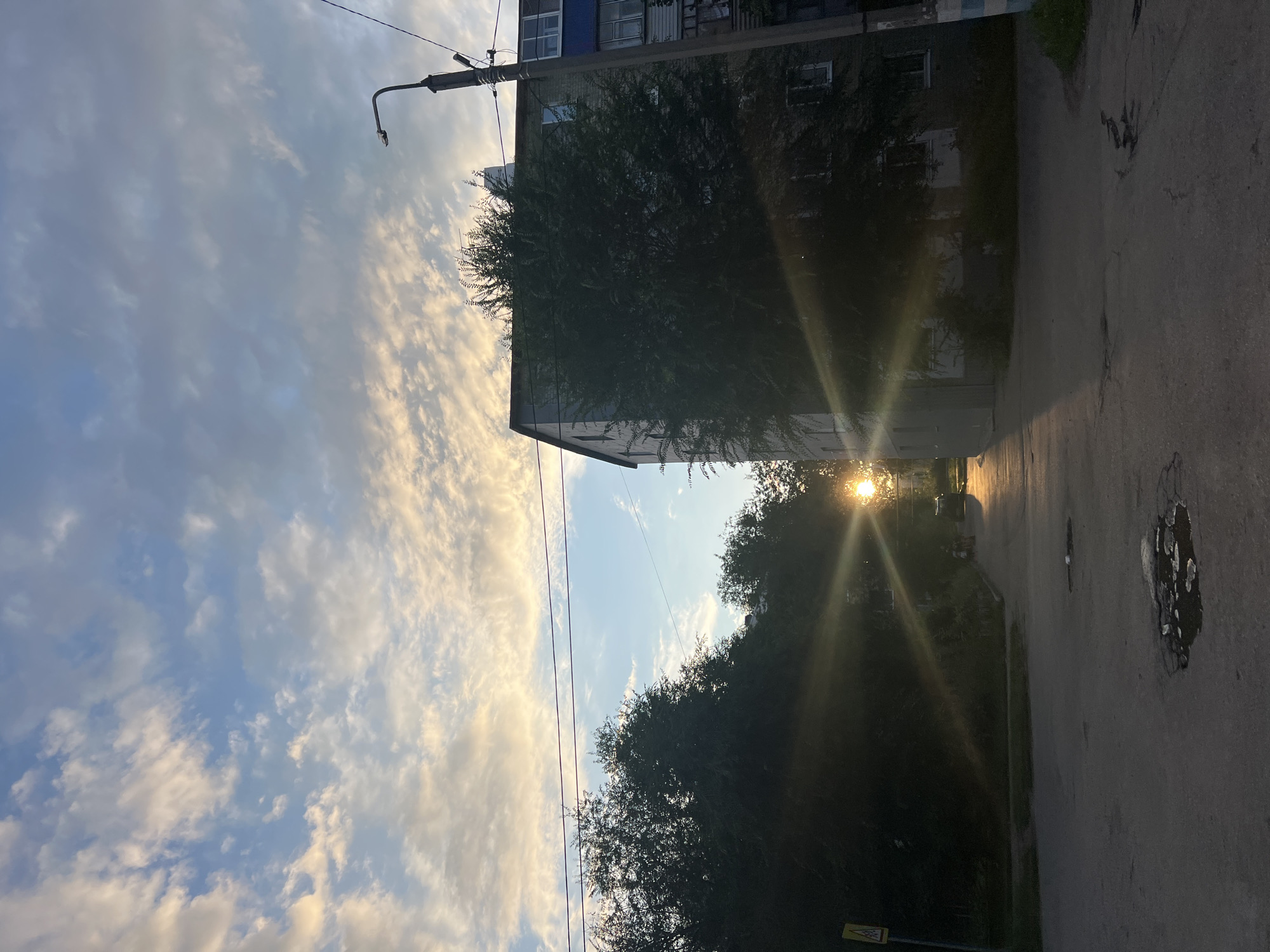 The photograph captures an urban street scene in Ulyanovsk, Russia, during a sunset. The sun, low in the sky, creates a dramatic lens flare that radiates outwards, illuminating the clouds and casting long shadows. In the foreground, a paved street shows signs of wear, with visible potholes filled with water. On the right side of the street, a multi-story residential building dominates the view, its facade partially obscured by a large, leafy tree. A street lamp stands tall next to the building. On the left side of the frame, more trees line the street, creating a darker, silhouetted area. A yellow road sign with a black symbol is visible near the left edge of the image. In the mid-ground, a few figures and a vehicle are present on the street, their forms softened by the low light and the sun's glare. The sky above is a mix of blue and scattered clouds, tinged with the warm hues of the setting sun.