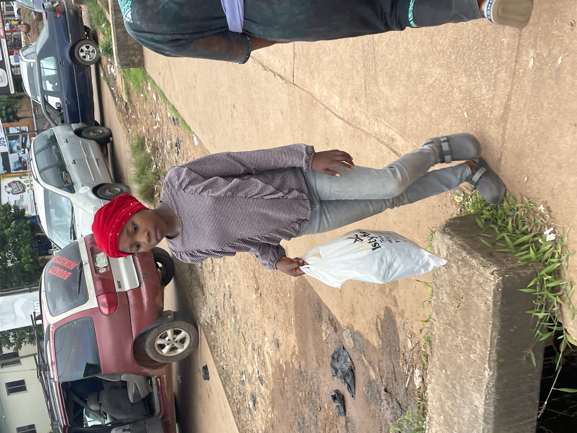 A young girl in a red headscarf and a checkered shirt walks along a sidewalk carrying a white plastic bag. She wears grey pants and dark clogs. Next to her, another person, partially visible, walks in the same direction. The sidewalk is next to a dirt road with a maroon minivan and other vehicles in the background. The environment appears to be an urban area in Jos, Nigeria, with buildings and commercial signage visible. The ground is littered with debris, including plastic bags.