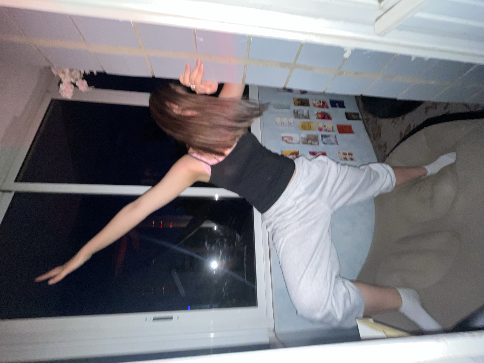 An individual is captured in an unusual, gravity-defying pose within what appears to be an indoor room at night. The subject, likely a young woman, has medium-brown hair that covers her face and is wearing a black tank top, light grey or white cropped pants, and white socks. She is positioned horizontally across the frame, with her left arm extended towards a large window. Her legs are bent, and her right hand is raised towards the top of the frame.
Through the window, a dark night sky with faint, blurry distant lights is visible, suggesting an exterior urban or developed area. Near the top of the window, a cluster of pink artificial flowers hangs down. The ceiling above the subject is tiled or paneled in a light color. On a wall behind the subject, numerous small, rectangular pictures or postcards are taped, forming a collage. Below the subject's legs, a patterned rug or carpet is visible on the floor. The scene suggests a playful or artistic photographic moment, utilizing perspective to create a surreal effect in a dimly lit personal space. No text is visible in the image.