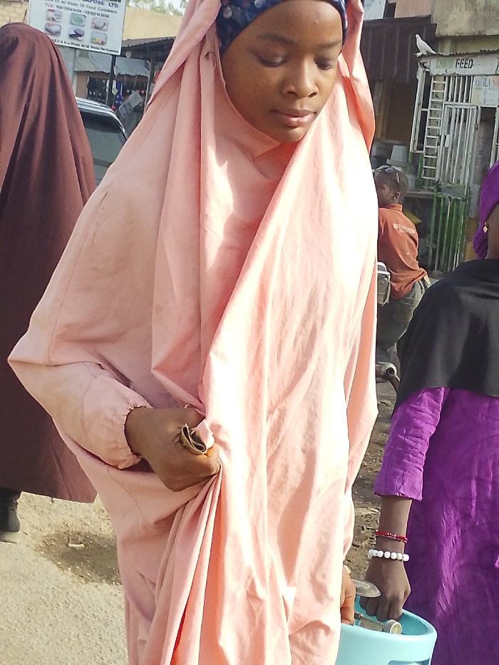 The image captures an outdoor street scene in Garun Gawa, Nigeria. The primary subject, a young woman of African descent, is centrally positioned, wearing a light pink, full-body garment over a patterned headscarf. Her gaze is directed downwards. Her right hand, adorned with a brass-colored ring on the index finger, grips the fabric of her attire. To her immediate right, a younger individual wears a vibrant purple long-sleeved top and a black head covering. This individual's left wrist displays red and white beaded bracelets, and they hold a light blue plastic container containing coins. In the far left background, a person partially obscured by a dark brown garment is visible next to a white vehicle. Further in the background, a person in an orange-brown shirt is partially visible, appearing to be near a motorcycle. Architectural elements include a commercial sign reading "PURPOSE LTD" and displaying household product images, and a structure with metal grates featuring a "FEED" sign, above which a white bird is perched. The ground surface is unpaved.